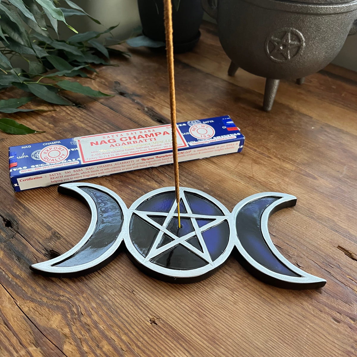 A triple moon pentacle incense holder with a central pentagram design sits on a wooden table, with a burning incense stick inserted into the center. A box of Nag Champa incense is visible in the background.