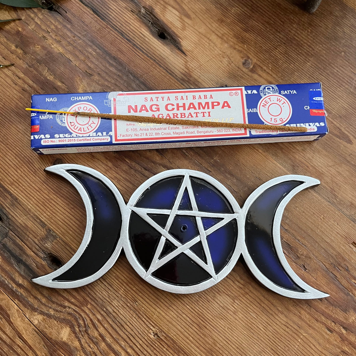 A silver and dark blue glass moon and pentacle incense holder displayed on a wooden surface, with a box of Satya Shiva Babi Nag Champa Agarbatti incense sticks placed behind them.