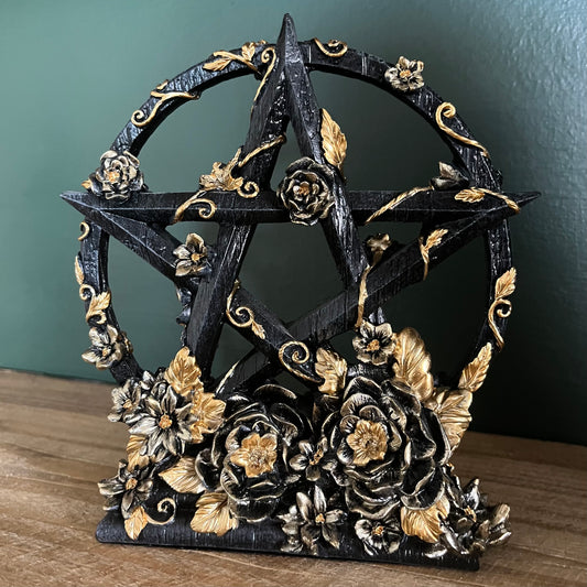 A black pentagram star sculpture decorated with gold and silver floral elements, displayed on a wooden surface against a dark green wall.