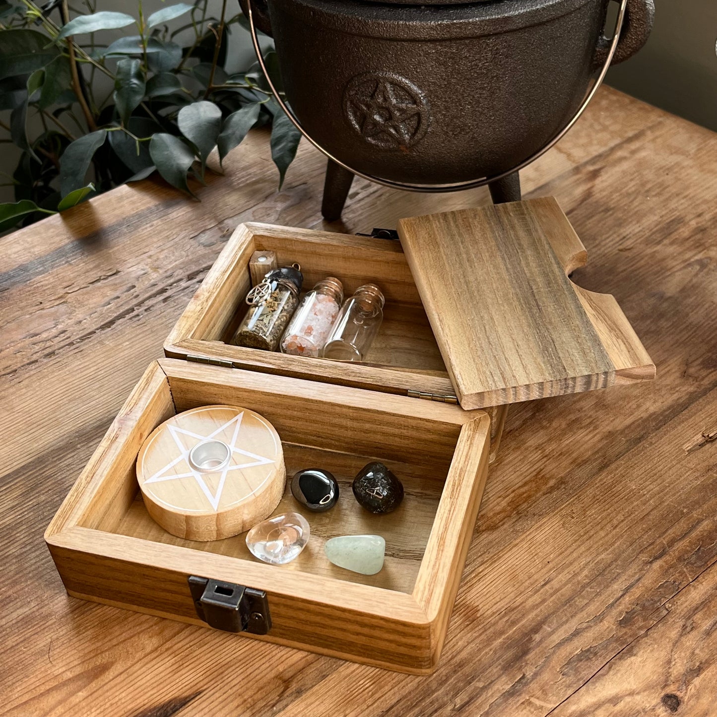 Inside the Wooden box with beautiful tree of life printed design on the lid, shows a separate magnetic compartment sat on a wooden table. The contents of the box can be seen. They are witchcraft or pagan tools. A white spell candle with a wooden candle holder with pentagram design. a palo santo smudge stick, 4 crystal tumble stones to represent the 4 elements, 3 spell bottles 1 containing herbs for protection, 1 containing salt for cleansing and 1 empty bottle.