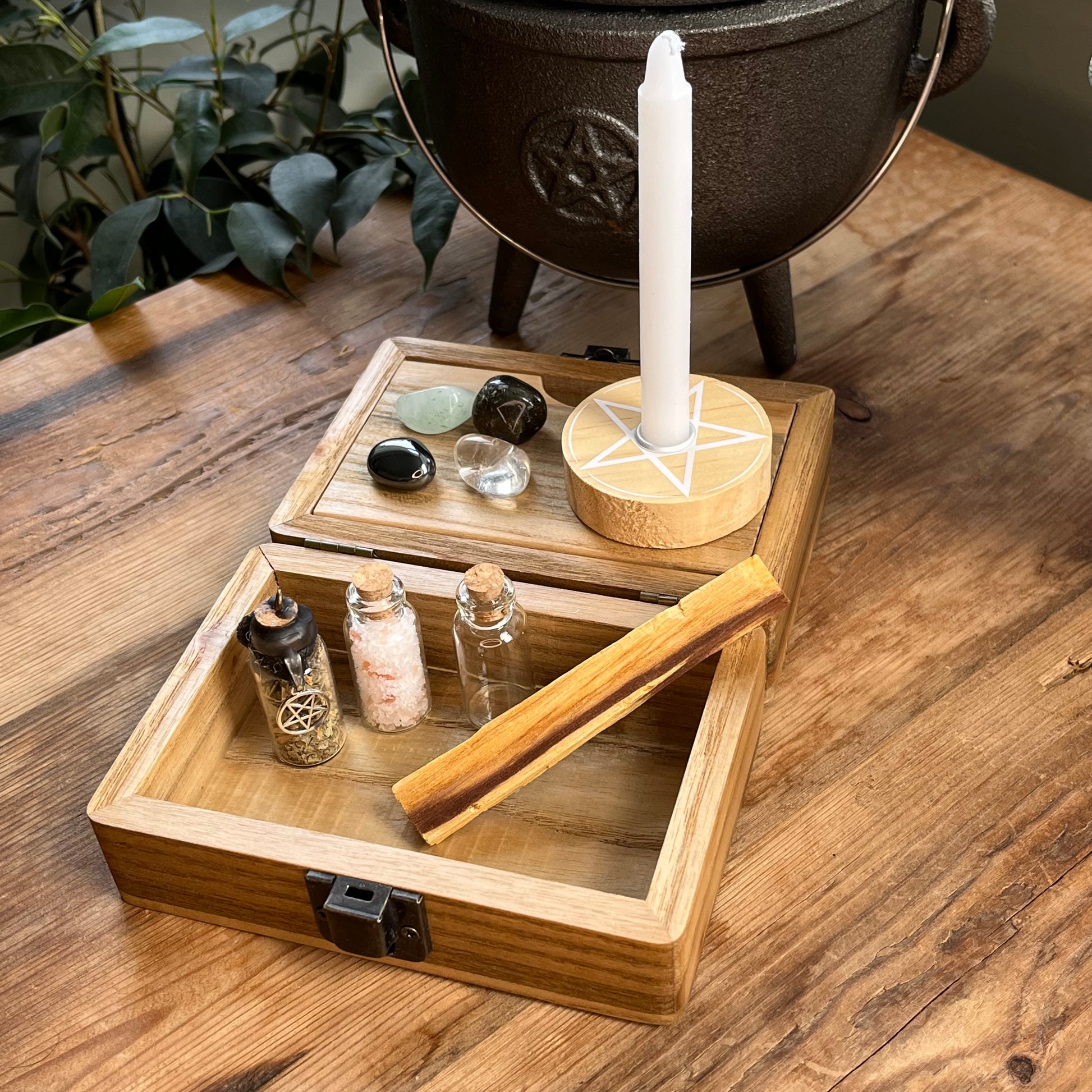 Inside the Wooden box with beautiful tree of life printed design on the lid, shows a separate magnetic compartment sat on a wooden table. The contents of the box can be seen. They are witchcraft or pagan tools. A white spell candle with a wooden candle holder with pentagram design. a palo santo smudge stick, 4 crystal tumble stones to represent the 4 elements, 3 spell bottles 1 containing herbs for protection, 1 containing salt for cleansing and 1 empty bottle.