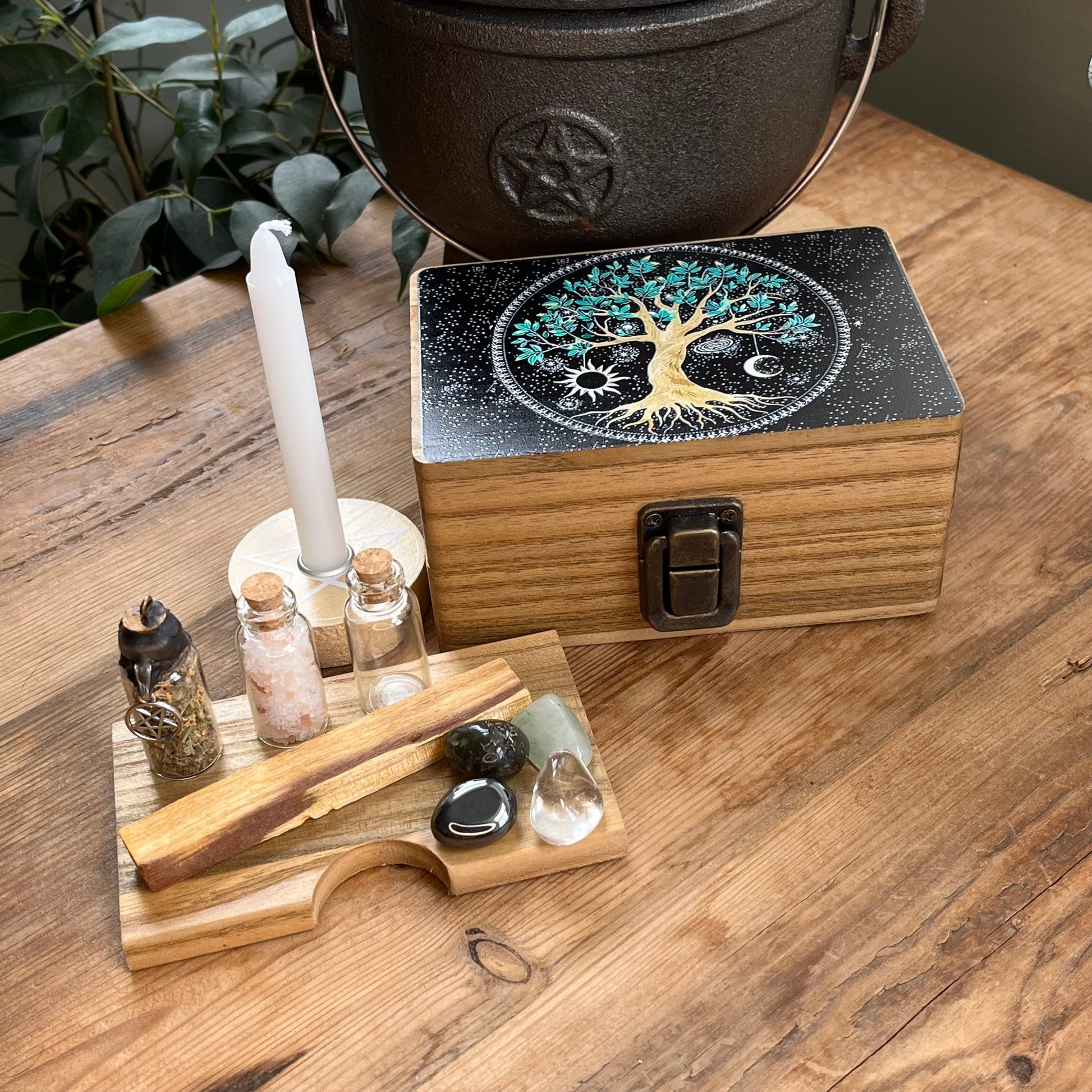 Wooden box with beautiful tree of life printed design on the lid on a table alongside the contents of the box, which are witchcraft or pagan tools. A white spell candle with a wooden candle holder with pentagram design. a palo santo smudge stick, 4 crystal tumble stones to represent the 4 elements, 3 spell bottles 1 containing herbs for protection, 1 containing salt for cleansing and 1 empty bottle.