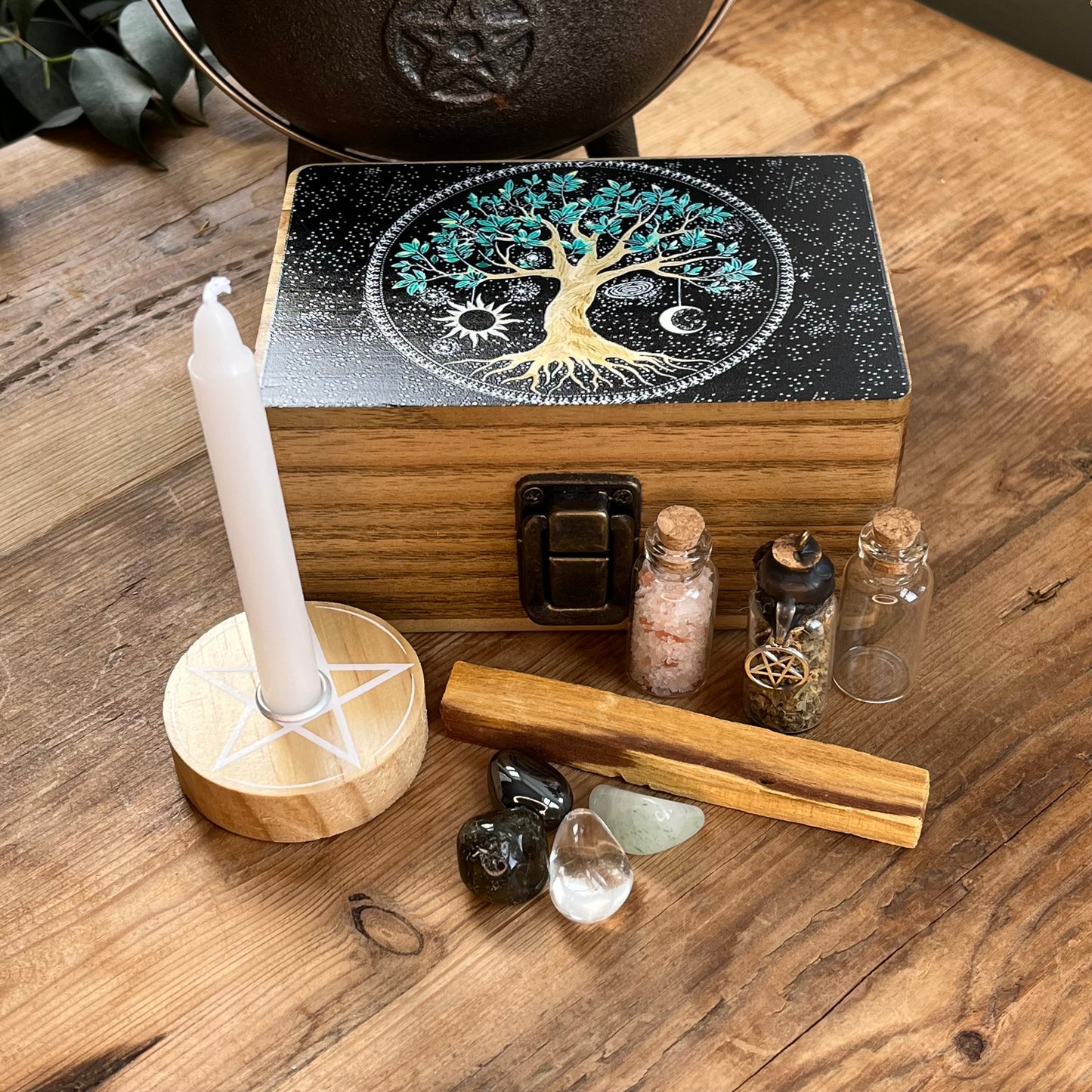 Wooden box with beautiful tree of life printed design on the lid on a table alongside the contents of the box, which are witchcraft or pagan tools. A white spell candle with a wooden candle holder with pentagram design. a palo santo smudge stick, 4 crystal tumble stones to represent the 4 elements, 3 spell bottles 1 containing herbs for protection, 1 containing salt for cleansing and 1 empty bottle.