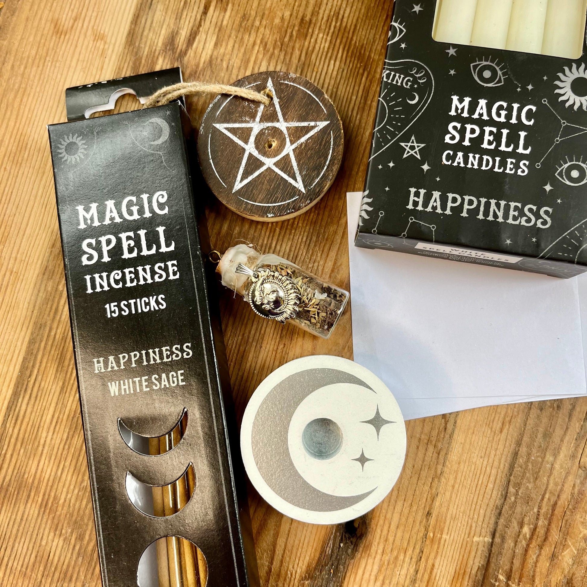 'Happiness Spell Kit' Contents displayed on a wooden table. Pack of Happiness white sage Incense sticks, and wooden pentagram incense holder, Pack of white magic spell candles, wooden white spell candle holder with crescent moon and star design, squares of white paper and a happiness spell bottle containing herbs and sealed with white wax and a charm.