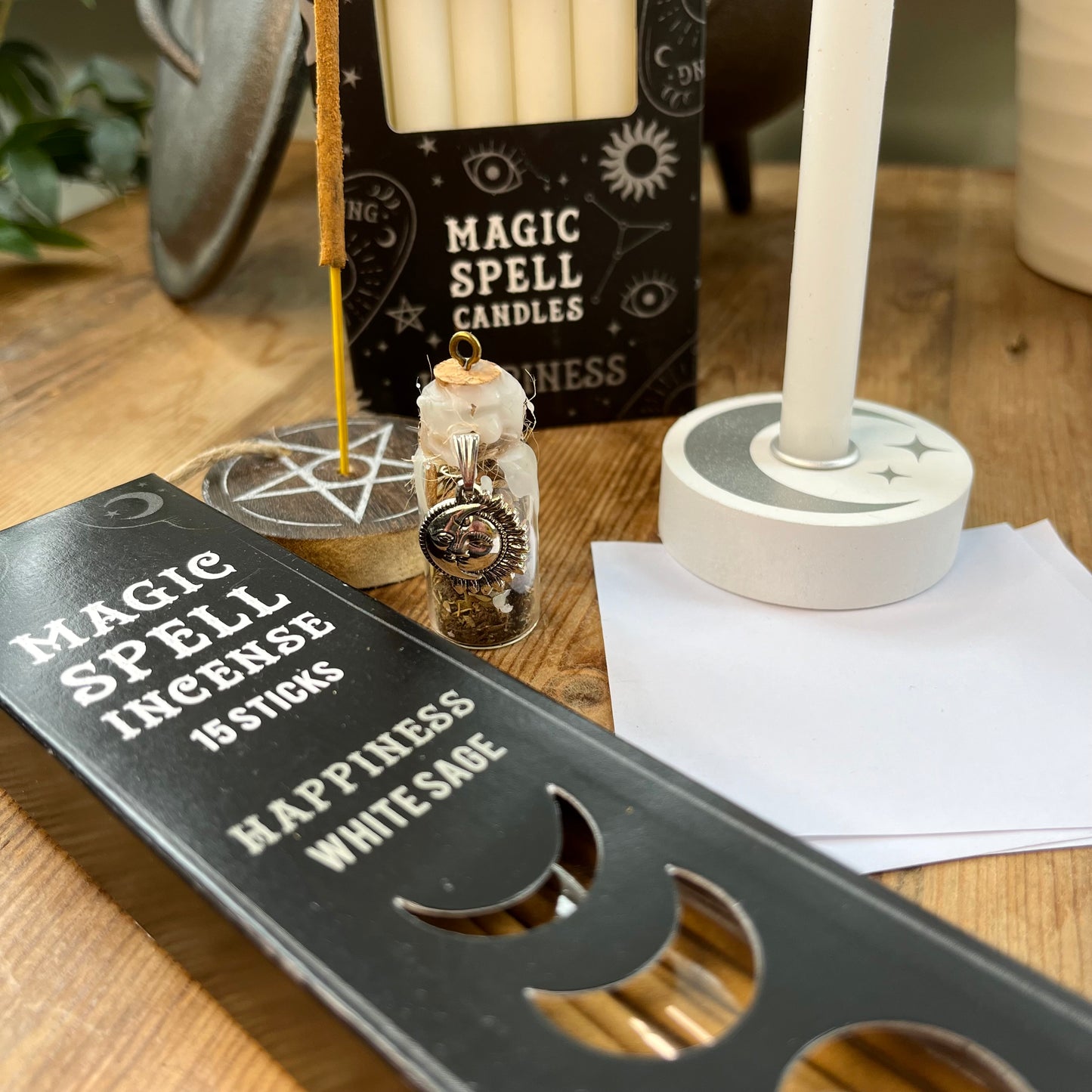 'Happiness Spell Kit' Contents displayed on a wooden table. Pack of Happiness white sage Incense sticks, and wooden pentagram incense holder, Pack of white magic spell candles, wooden white spell candle holder with crescent moon and star design, squares of white paper and a happiness spell bottle containing herbs and sealed with white wax and a charm.