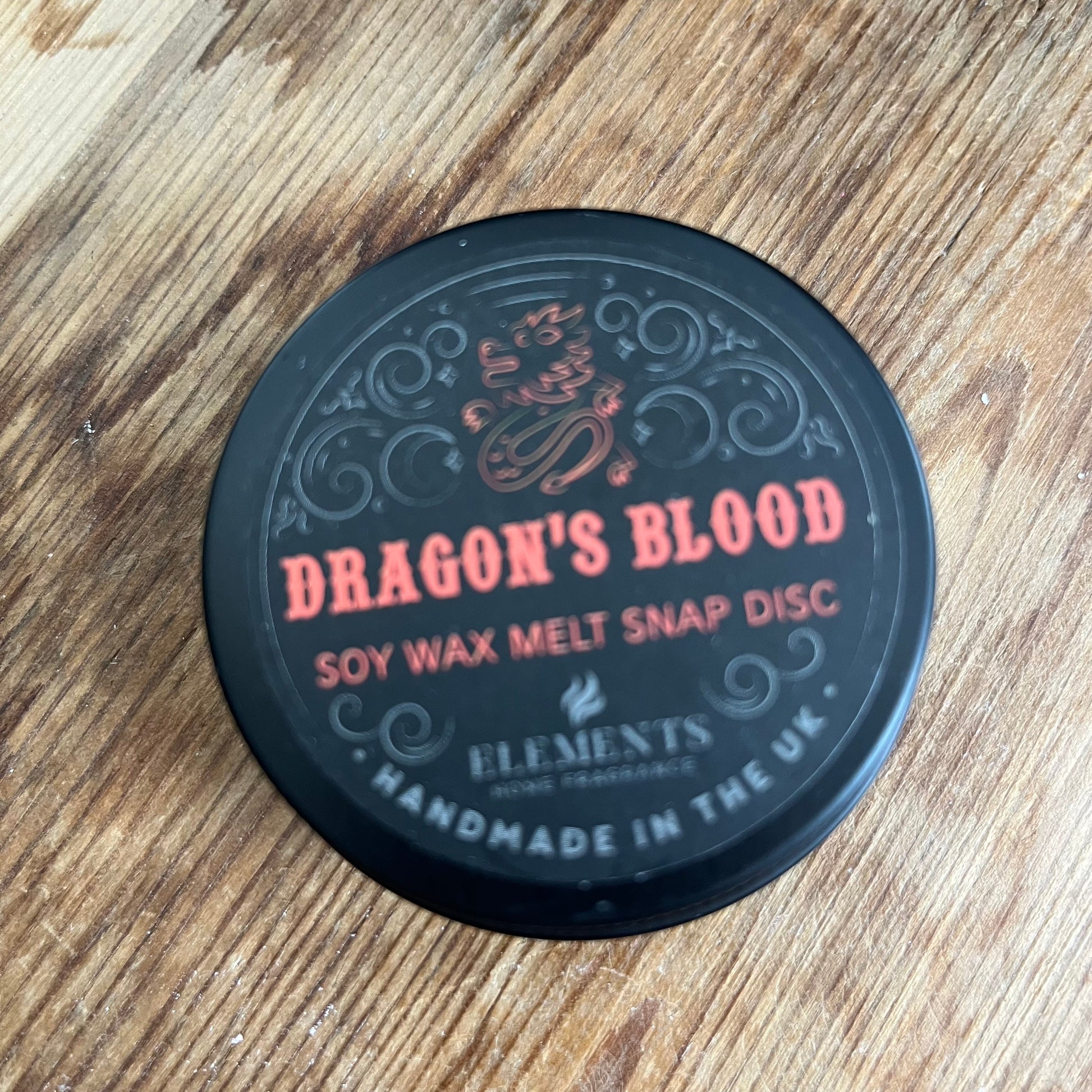 A circular tin containing a red wax melt divided into 4 sections. The tin lid reads Dragons blood soy wax melt snap disc 