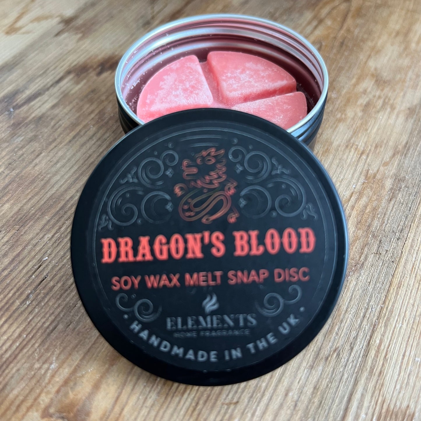 A circular tin containing a red wax melt divided into 4 sections. The tin lid reads Dragons blood soy wax melt snap disc 