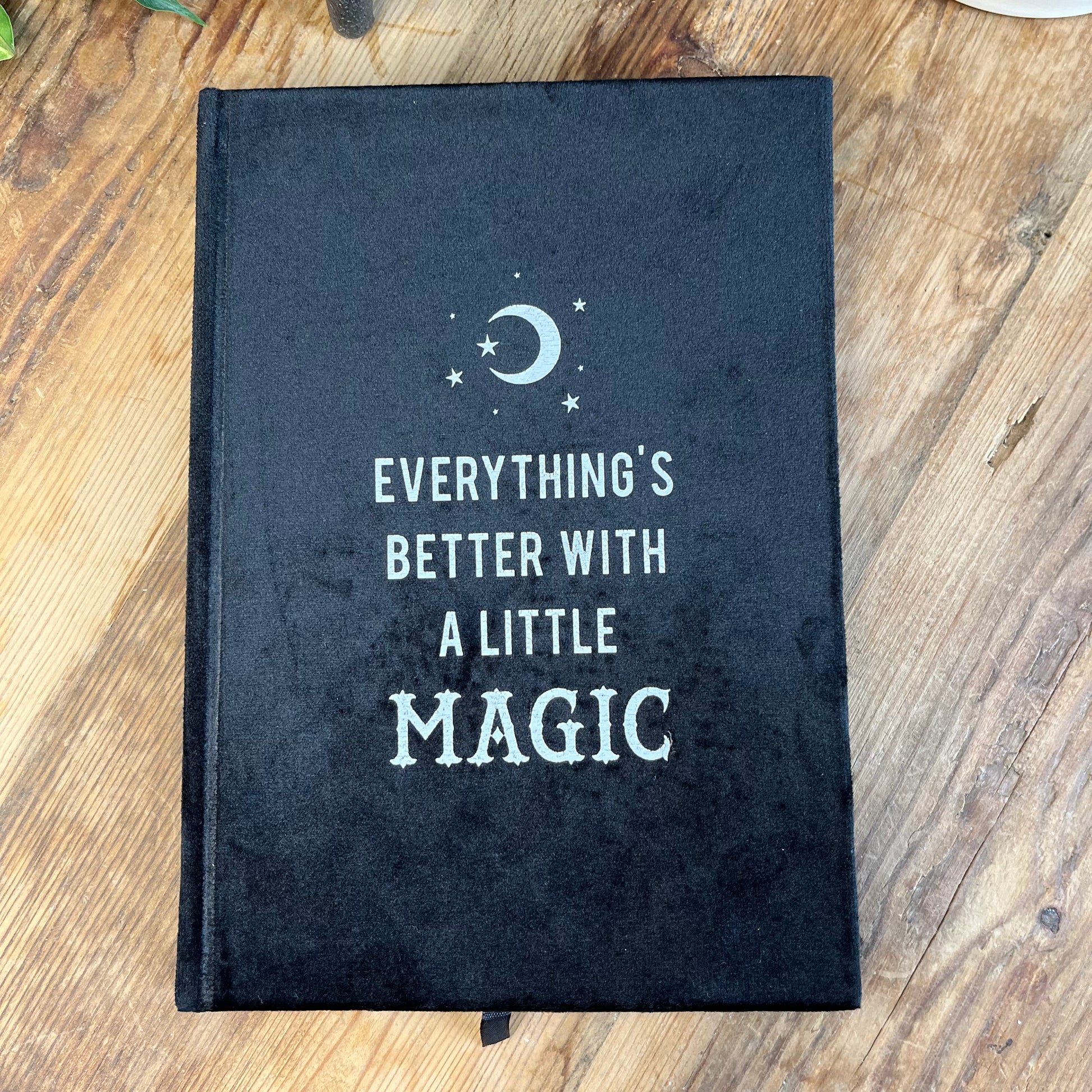 Black velvet notebook. Front cover has moon and stars design and the words 'everything's better with a little magic' The notebook is pictured with a second notebook that is open and pictured with a pen and witchy stickers 