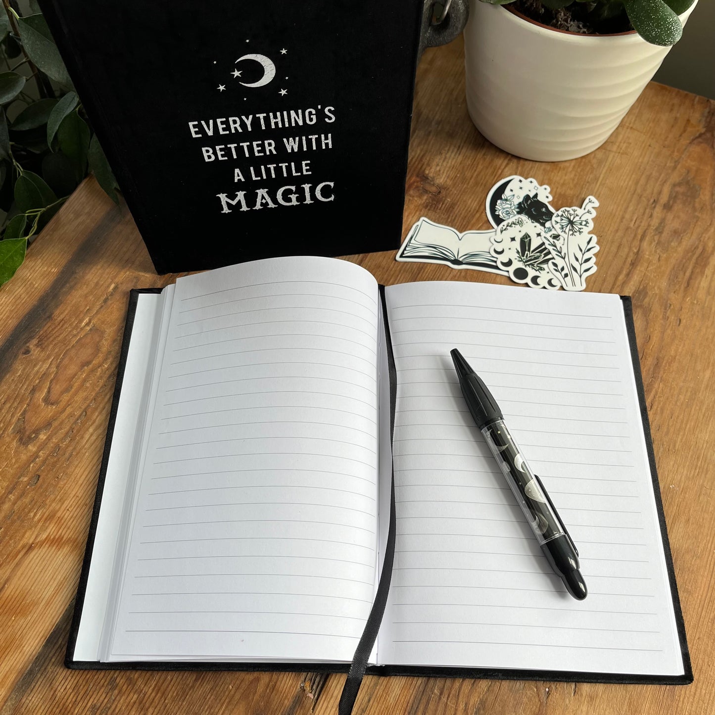 Black velvet notebook. Front cover has moon and stars design and the words 'everything's better with a little magic' The notebook is pictured with a second notebook that is open and pictured with a pen and witchy stickers 