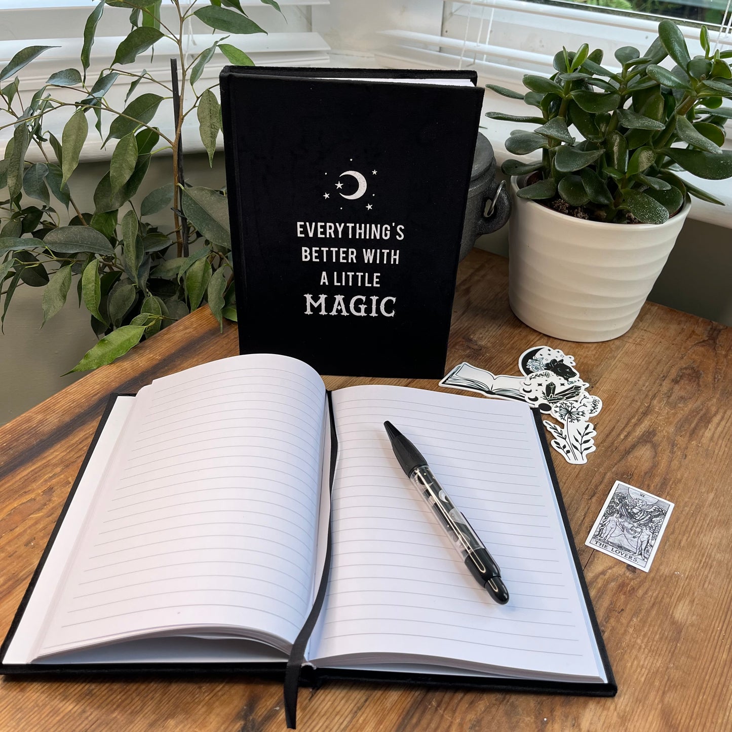 Black velvet notebook. Front cover has moon and stars design and the words 'everything's better with a little magic' The notebook is pictured with a second notebook that is open and pictured with a pen and witchy stickers 