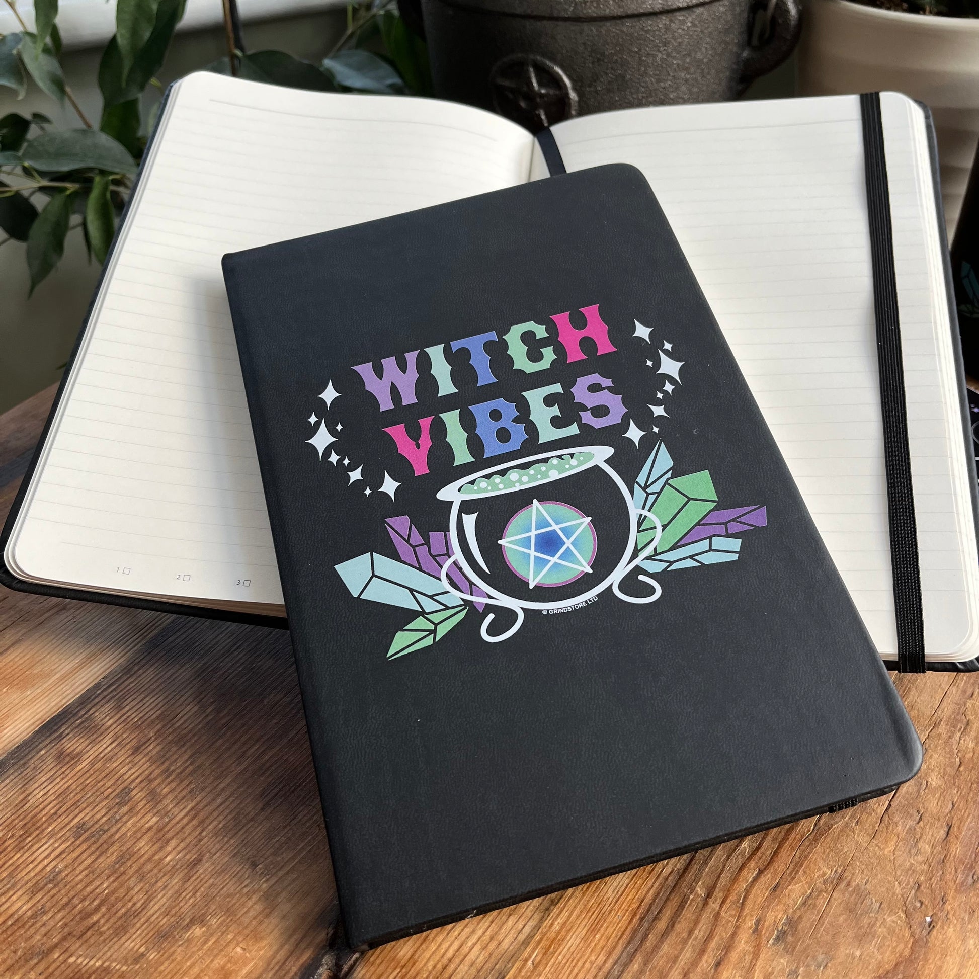 Black hard cover notebook. Front cover  has a graphic of a cauldron, crystals and a pentagram and the words 'witch vibes ' in shades of purple, green, blue and pink.  Image shows a closed notebook and an open notebook where the lined pages can be seen. 