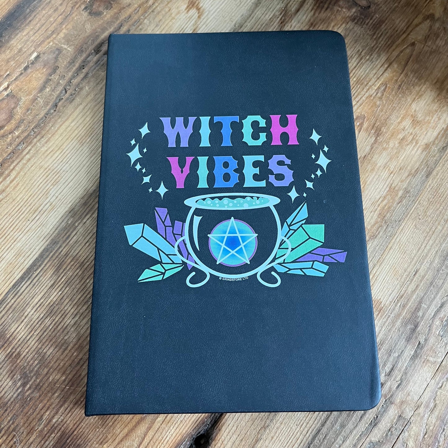 Black hard cover notebook. Front cover  has a graphic of a cauldron, crystals and a pentagram and the words 'witch vibes ' in shades of purple, green, blue and pink.  