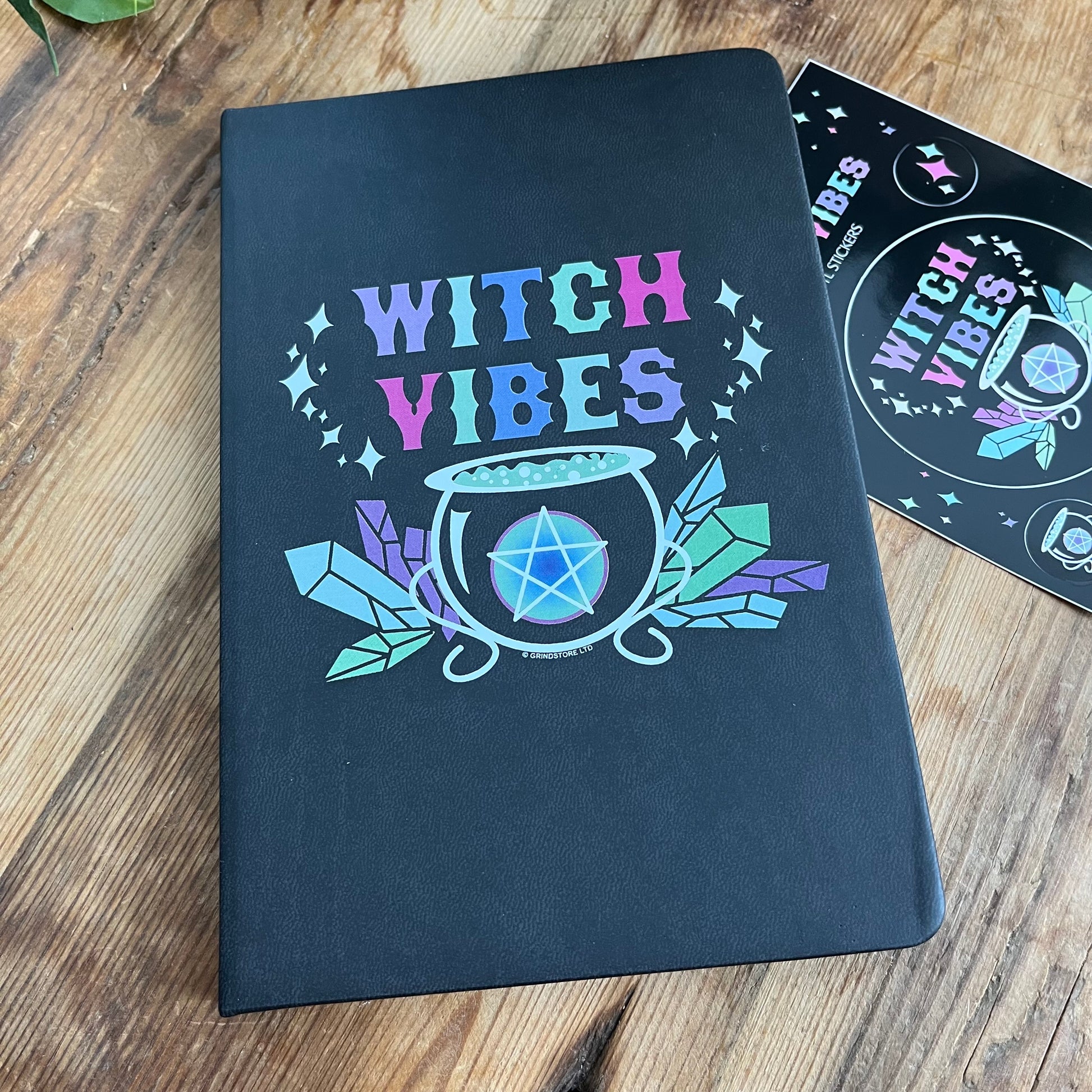 Black hard cover notebook. Front cover  has a graphic of a cauldron, crystals and a pentagram and the words 'witch vibes ' in shades of purple, green, blue and pink.  