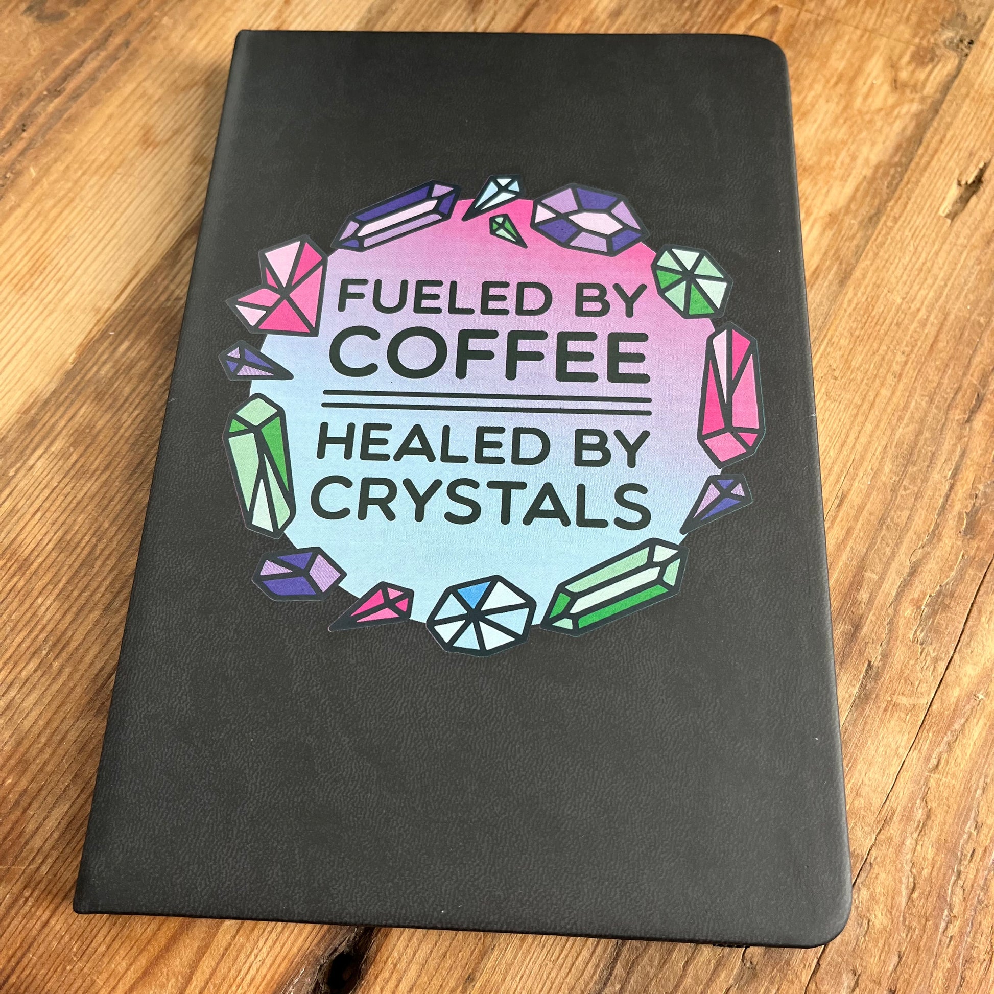 Black hard cover notebook with a graphic on the cover showing crystal shapes in shades of pink, blue and green and the words fueled by coffee, healed by crystals. 