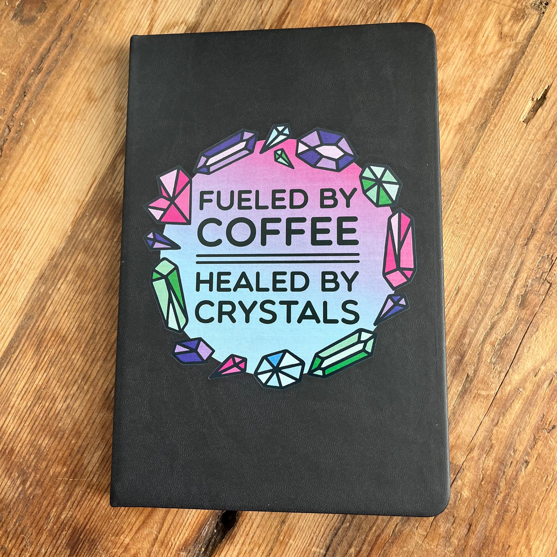 Black hard cover notebook with a graphic on the cover showing crystal shapes in shades of pink, blue and green and the words fueled by coffee, healed by crystals. 