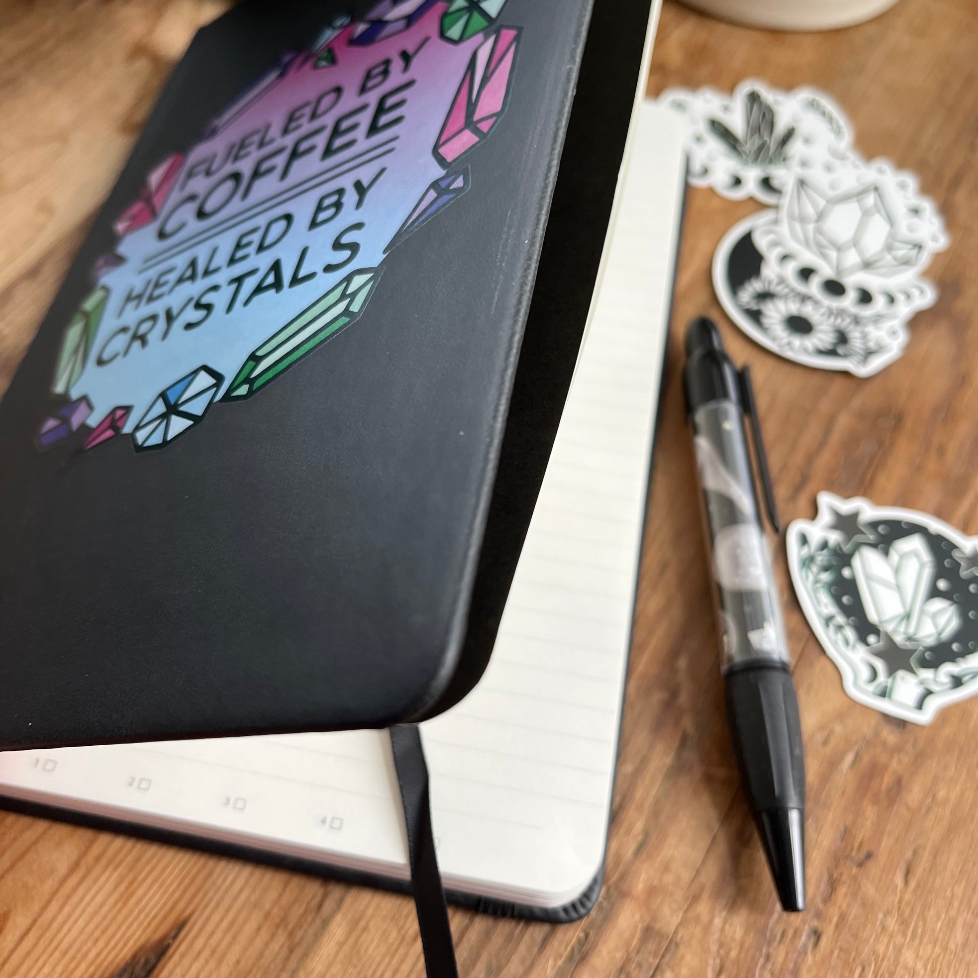 Black hard cover notebook with a graphic on the cover showing crystal shapes in shades of pink, blue and green and the words fueled by coffee, healed by crystals.  The notebook is held slightly open and the lined pages can be seen 