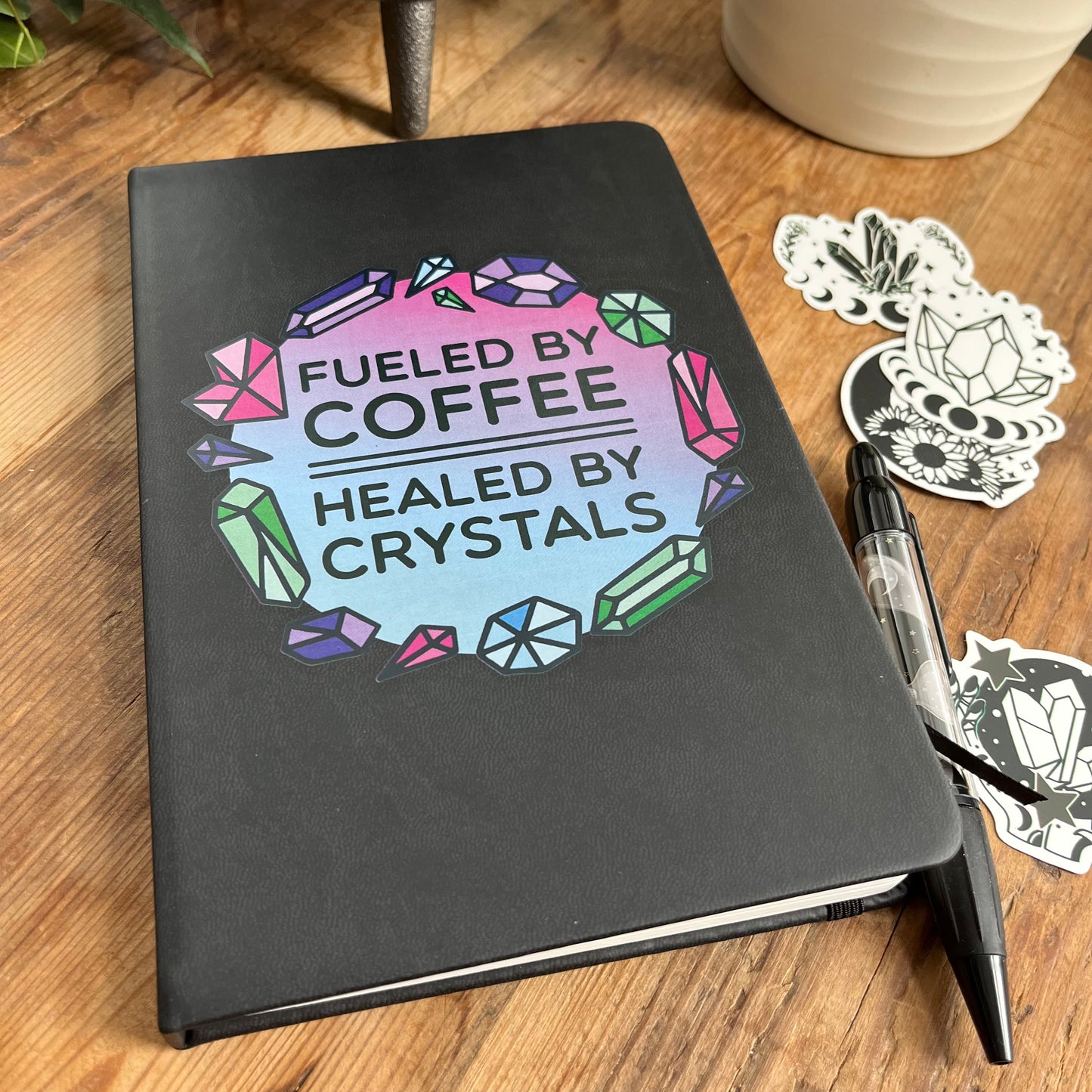 Black hard cover notebook with a graphic on the cover showing crystal shapes in shades of pink, blue and green and the words fueled by coffee, healed by crystals. 