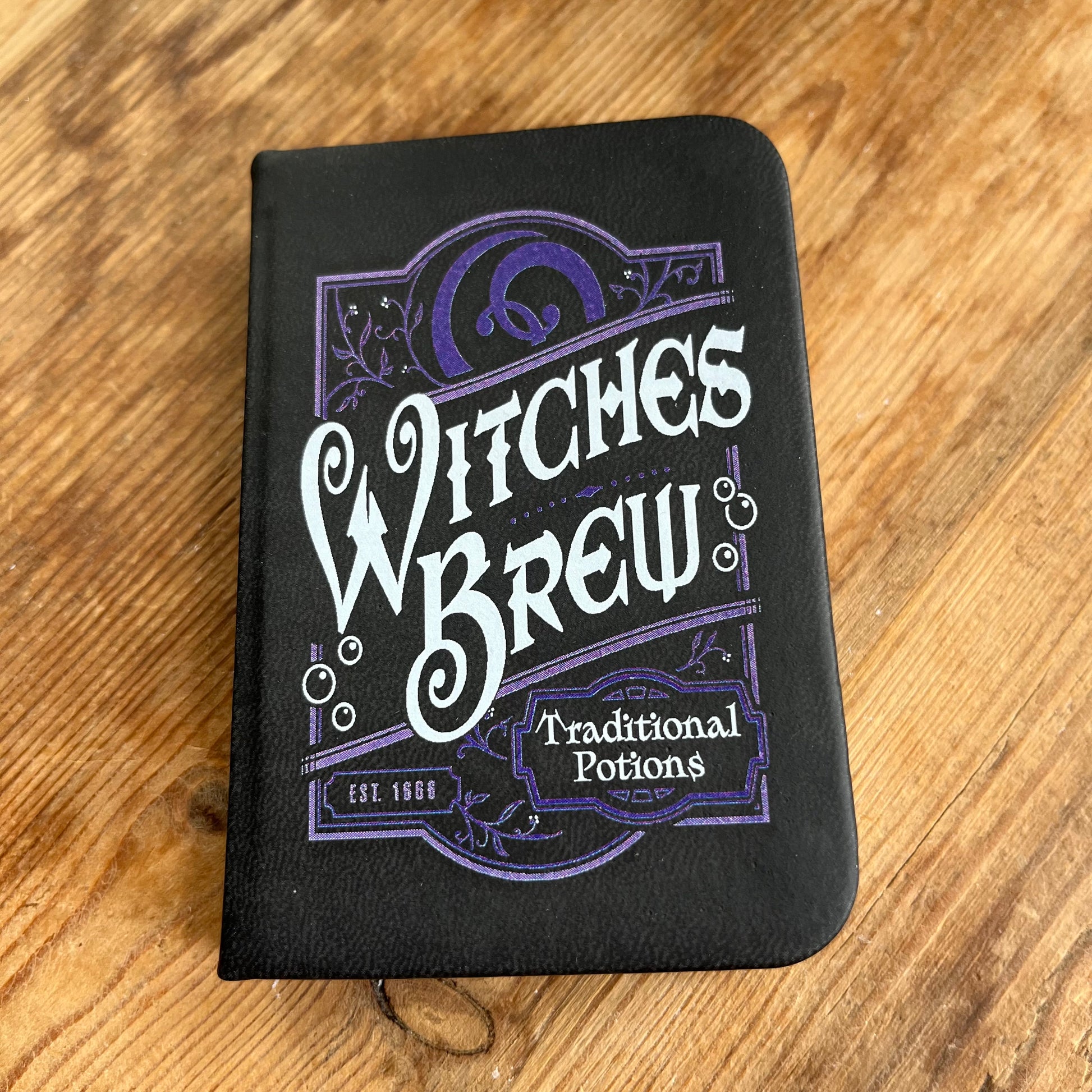 A small black pocket sized notebook with a purple pattern design and the words 'witches brew 'and 'traditional potions' in a curvy font on the front cover 