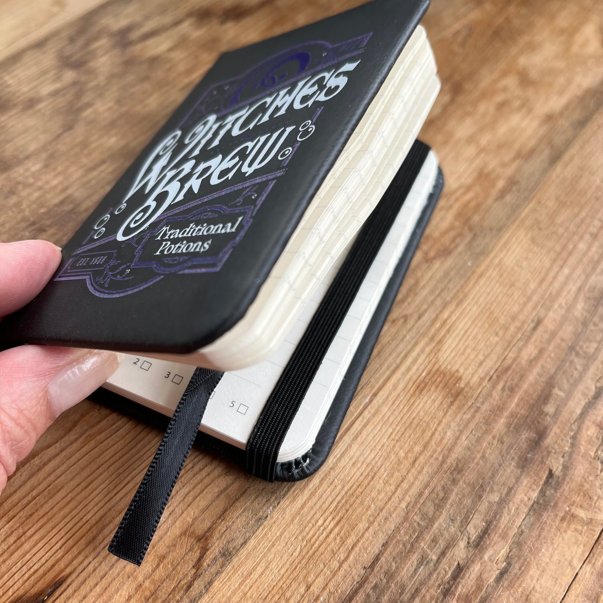 A small black pocket sized notebook with a purple pattern design and the words 'witches brew 'and 'traditional potions' in a curvy font on the front cover. the notebook is slightly open showing lined pages and an elastic book closure and ribbon page divider 