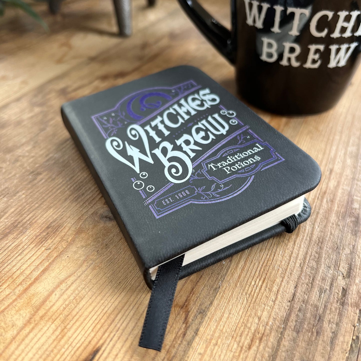 A small black pocket sized notebook with a purple pattern design and the words 'witches brew 'and 'traditional potions' in a curvy font on the front cover. In the background a mug also featuring the words witches brew can be seen 