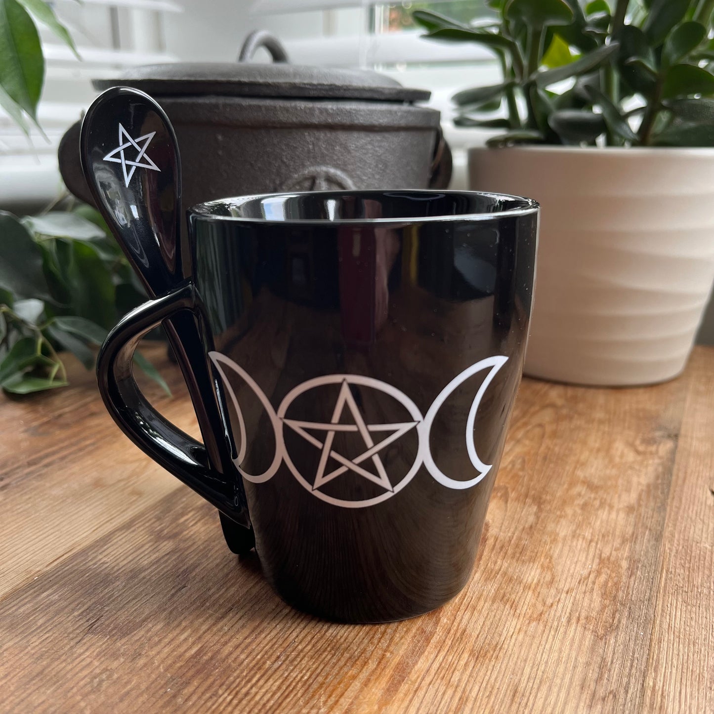 Black mug with a white triple moon and pentagram design on both sides. A matching ceramic poon slots into the handle of the mug 