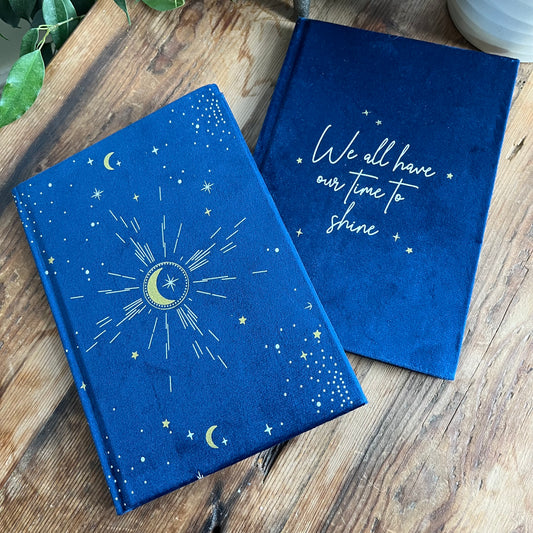 Two Blue velvet A5 notebooks sit on a wooden table. one notebook is face up showing the features words 'we all have our time to shine' The second notebook shows the back cover which features a moon and star design in gold 