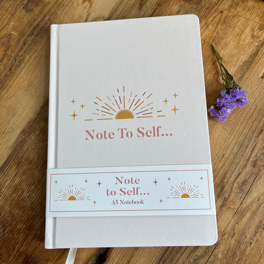 Very pale pink notebook featuring the words 'Note to Self' in pink and a golden sunrise sits on a wooden table next to a purple flower