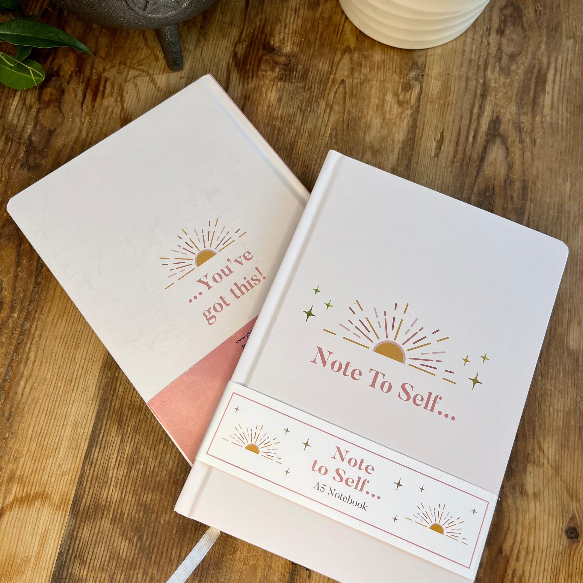 Pale pink notebooks one face up showing the front cover and the words 'note to self and gold sunset design. one face down showing the same sunset illustration and the words 'you've got this' 