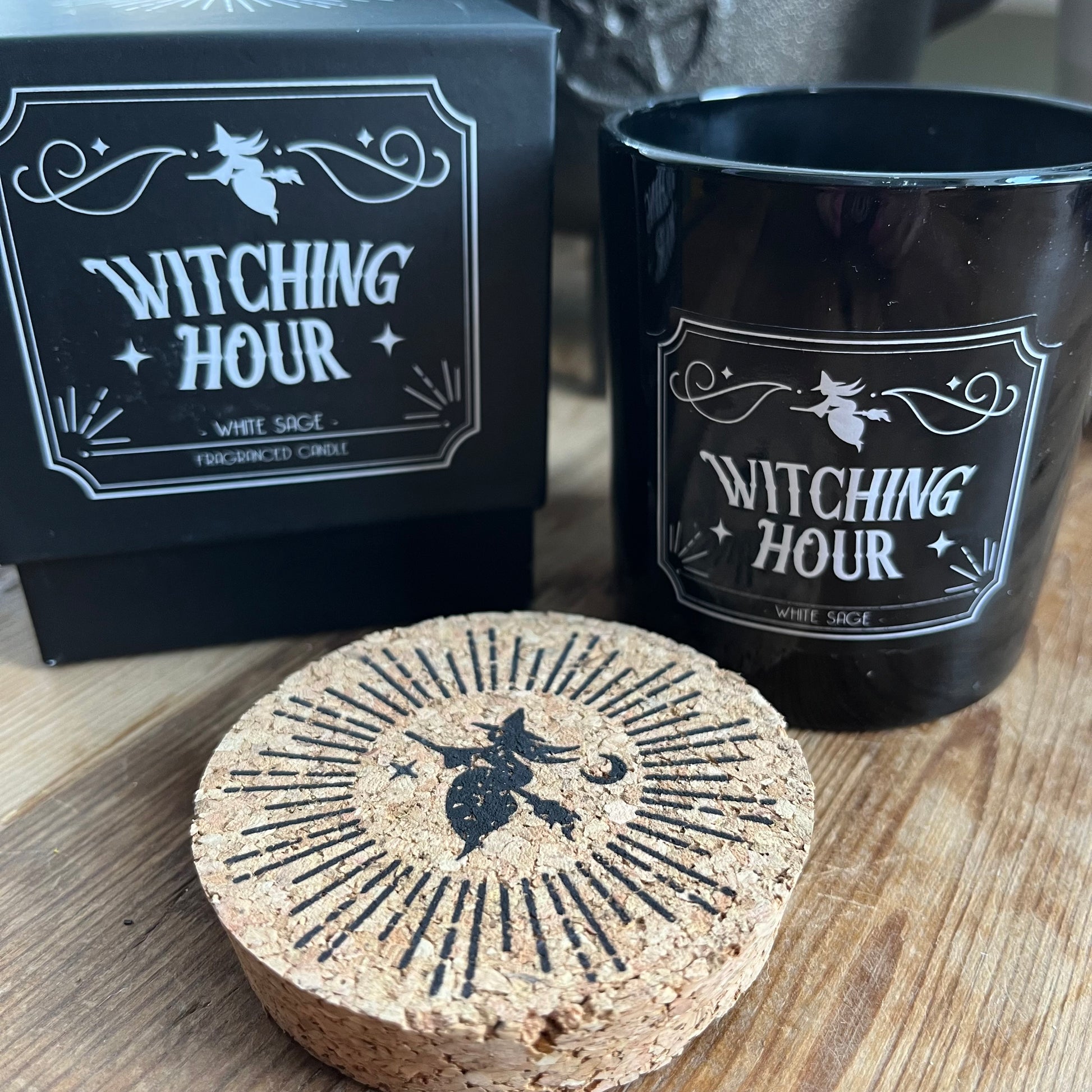 Boxed candle. The box is Black square with a lift off lid. It features a witch on a broomstick illustration and the words 'Witching Hour' 'White Sage fragranced candle.' The candle itself sits alongside the box. The candle holder is black glass and has the same words and illustration as the candle box, it also has a cork lid with the matching illustration on top in black.