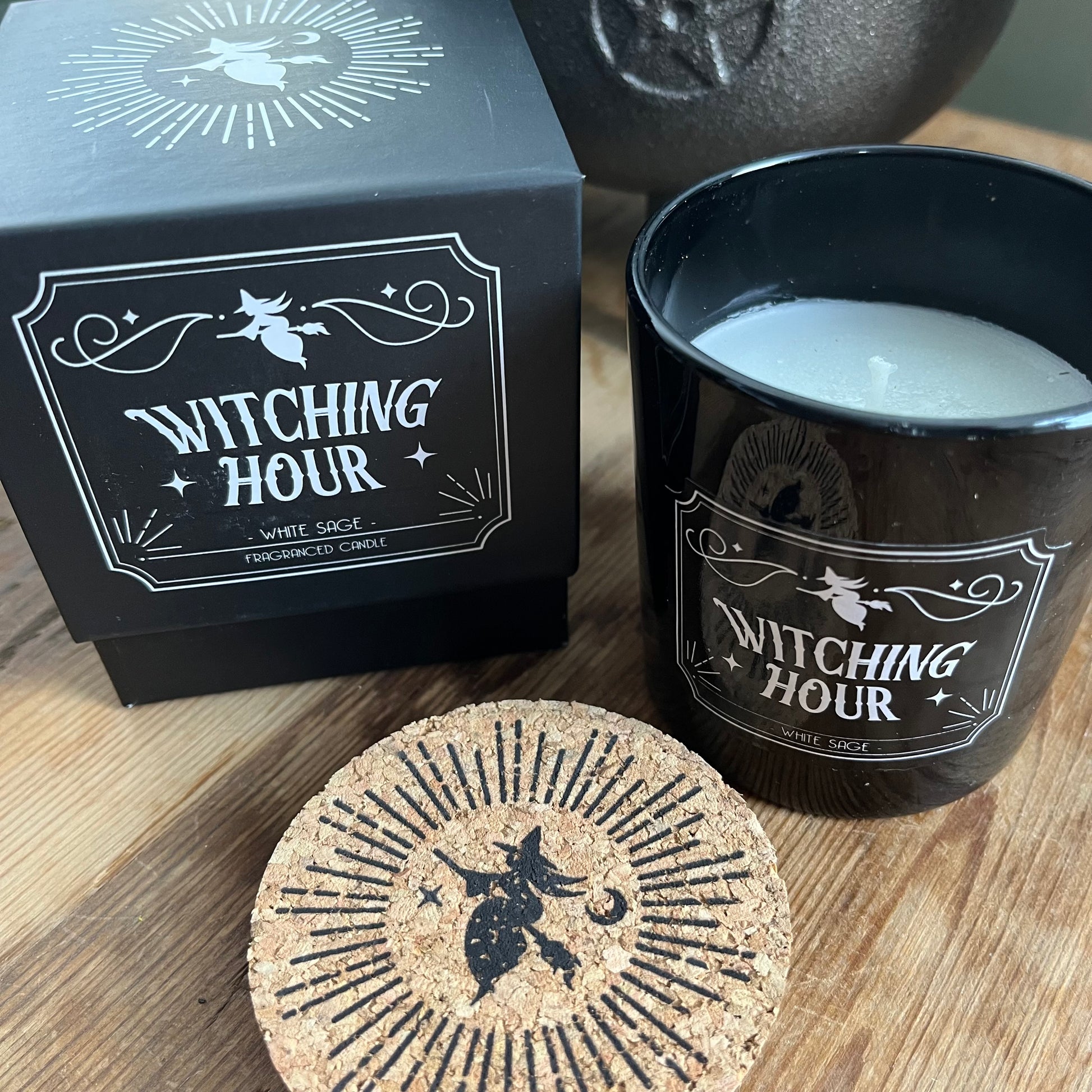 Boxed candle. The box is Black square with a lift off lid. It features a witch on a broomstick illustration and the words 'Witching Hour' 'White Sage fragranced candle.' The candle itself sits alongside the box. The candle holder is black glass and has the same words and illustration as the candle box, it also has a cork lid with the matching illustration on top in black.