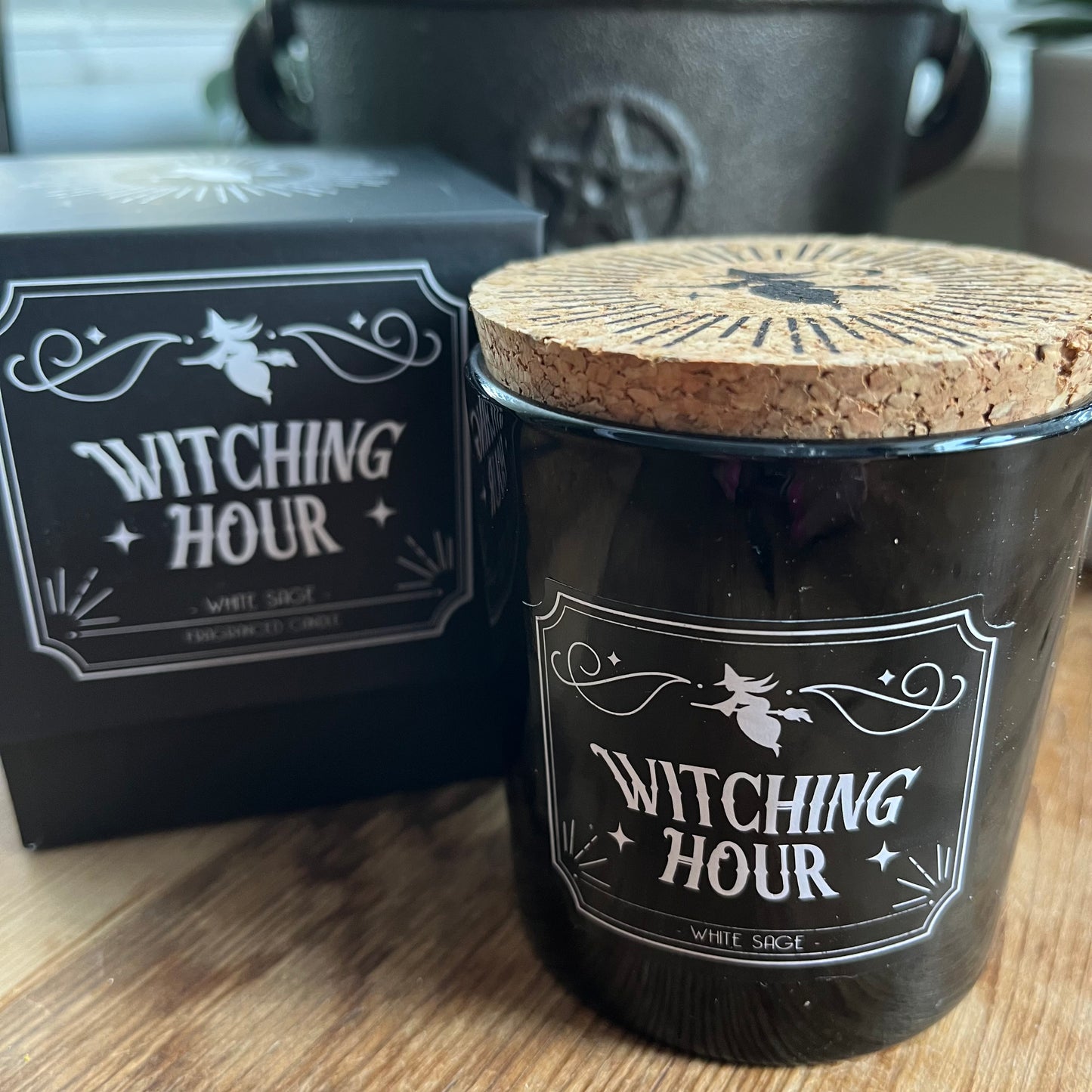 Boxed candle. The box is Black square with a lift off lid. It features a witch on a broomstick illustration and the words 'Witching Hour' 'White Sage fragranced candle.' The candle itself sits alongside the box. The candle holder is black glass and has the same words and illustration as the candle box, it also has a cork lid with the matching illustration on top in black.