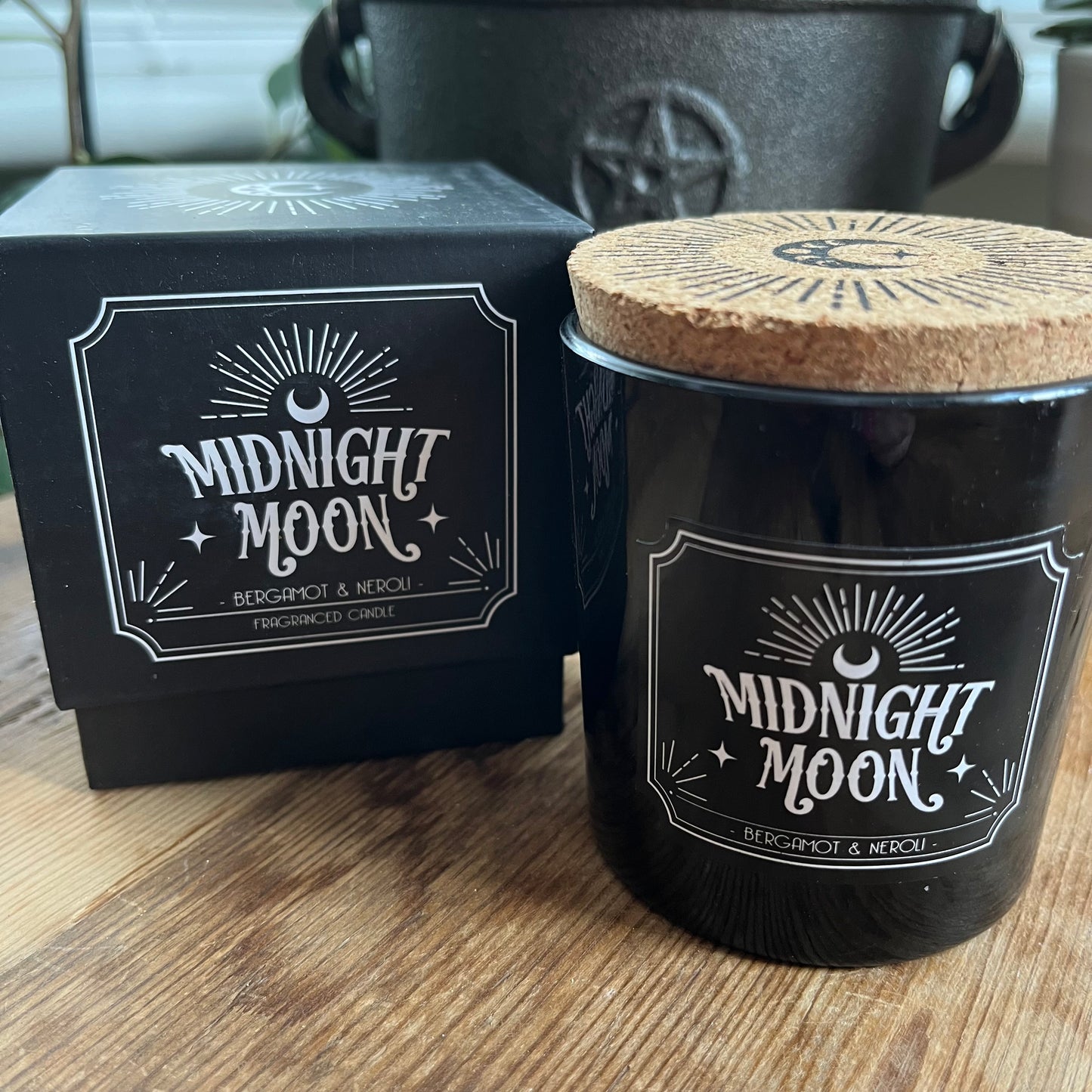 Boxed candle. The box is Black square with a lift off lid .It features a radiating crescent moon illustration and the words 'Midnight Moon' 'Bergamot and Neroli fragranced candle. The candle itself sits alongside the box. The candle holder is black glass and has the same words and illustration as the candle box, it also has a cork lid with the matching illustration on top in black.