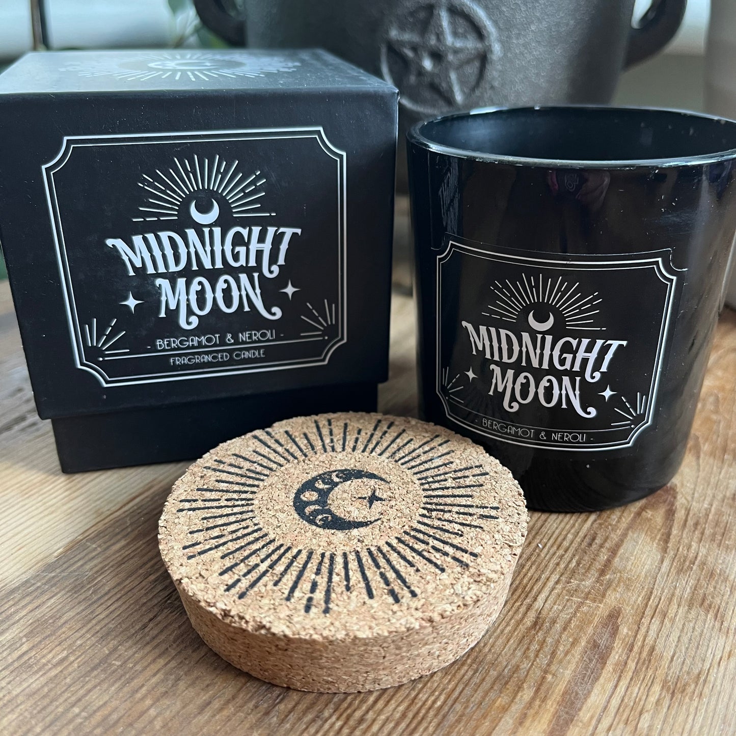 Boxed candle. The box is Black square with a lift off lid. It features a radiating crescent moon illustration and the words 'Midnight Moon' 'Bergamot and Neroli fragranced candle. The candle itself sits alongside the box. The candle holder is black glass and has the same words and illustration as the candle box, it also has a cork lid with the matching illustration on top in black.