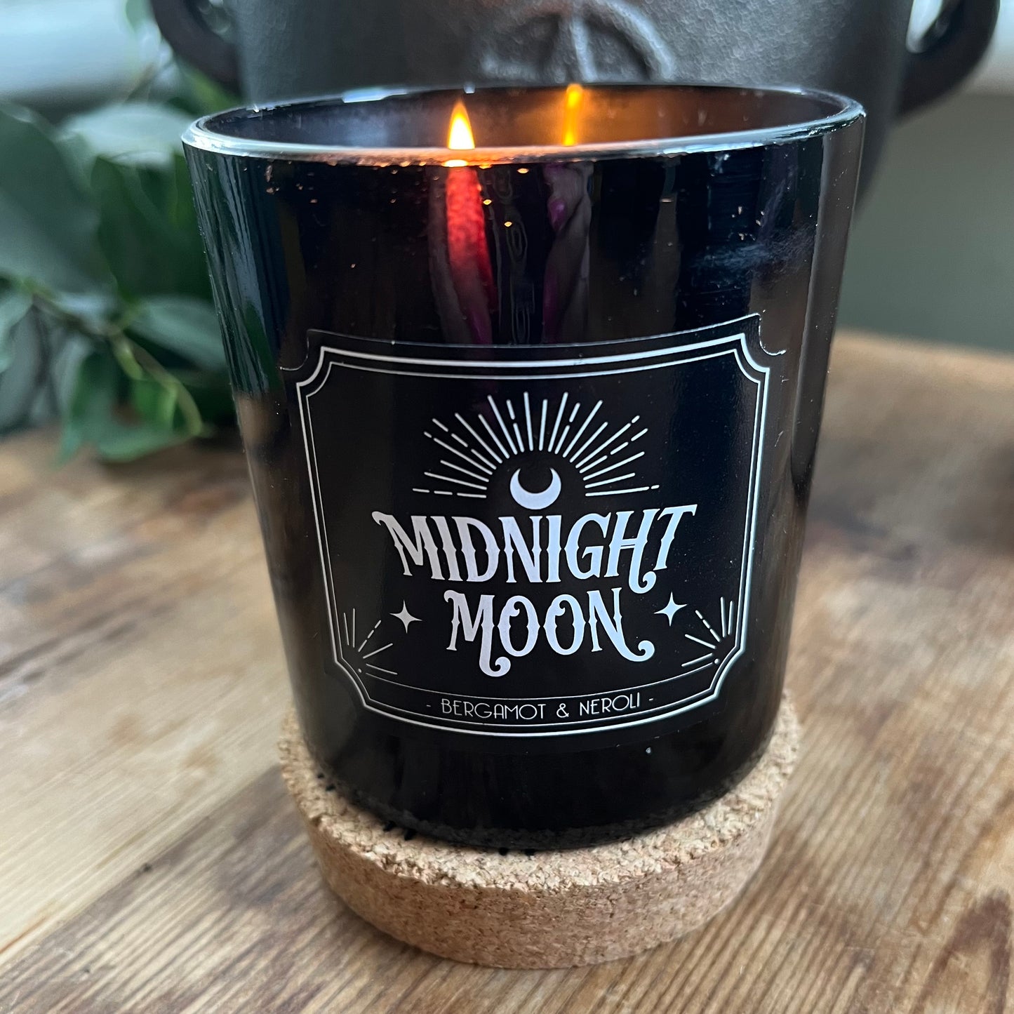 A lit candle in a black glass candle holder featuring a radiating crescent moon illustration and the words 'Midnight Moon' 'Bergamot and Neroli fragranced candle' on its side. The candle itself sits on it's cork lid.