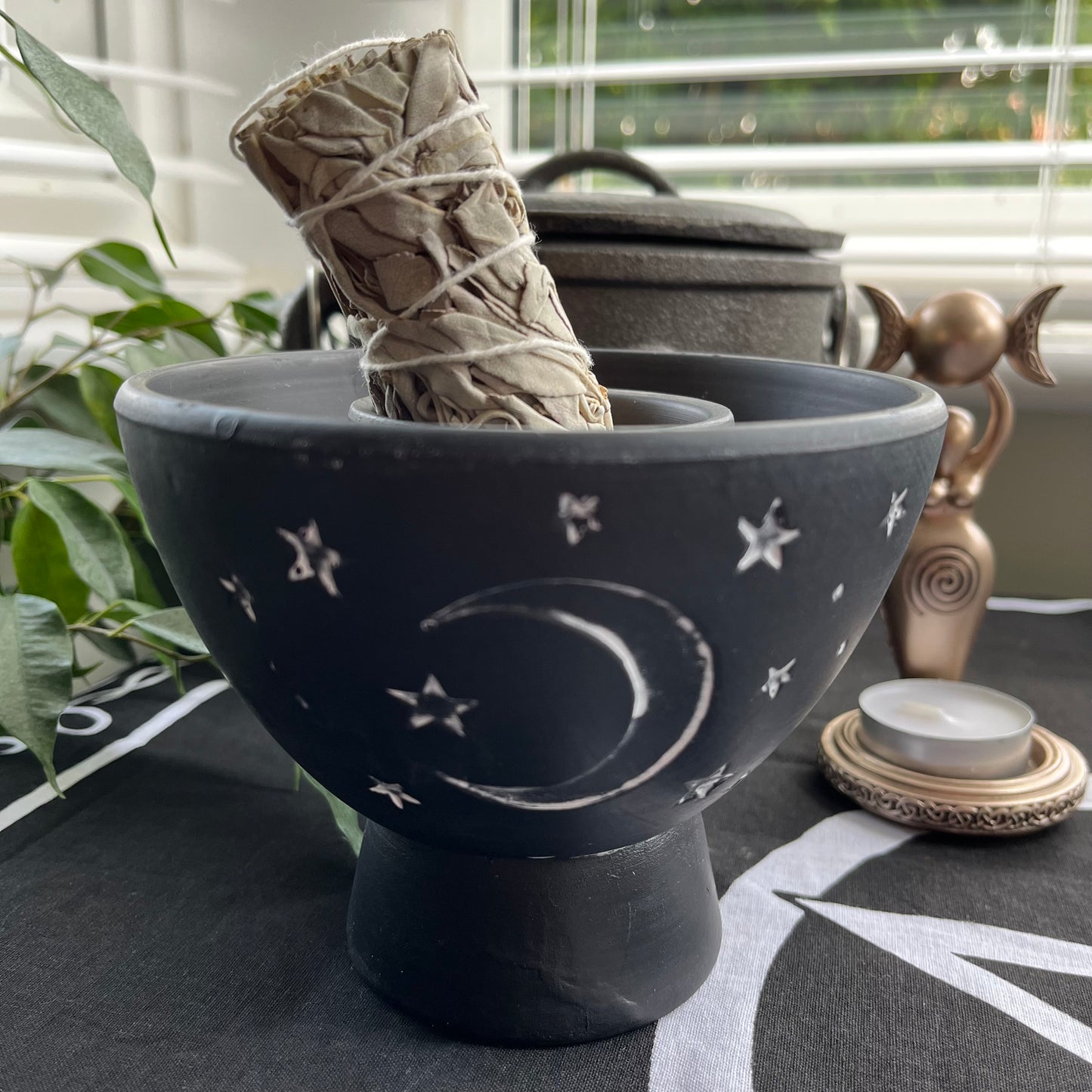 Rustic Black Terracota bowl designed to hold herbs and smudge sticks while burning. Has a white painted moon and stars motif on the side. Photographed on an altar with a smudge stick