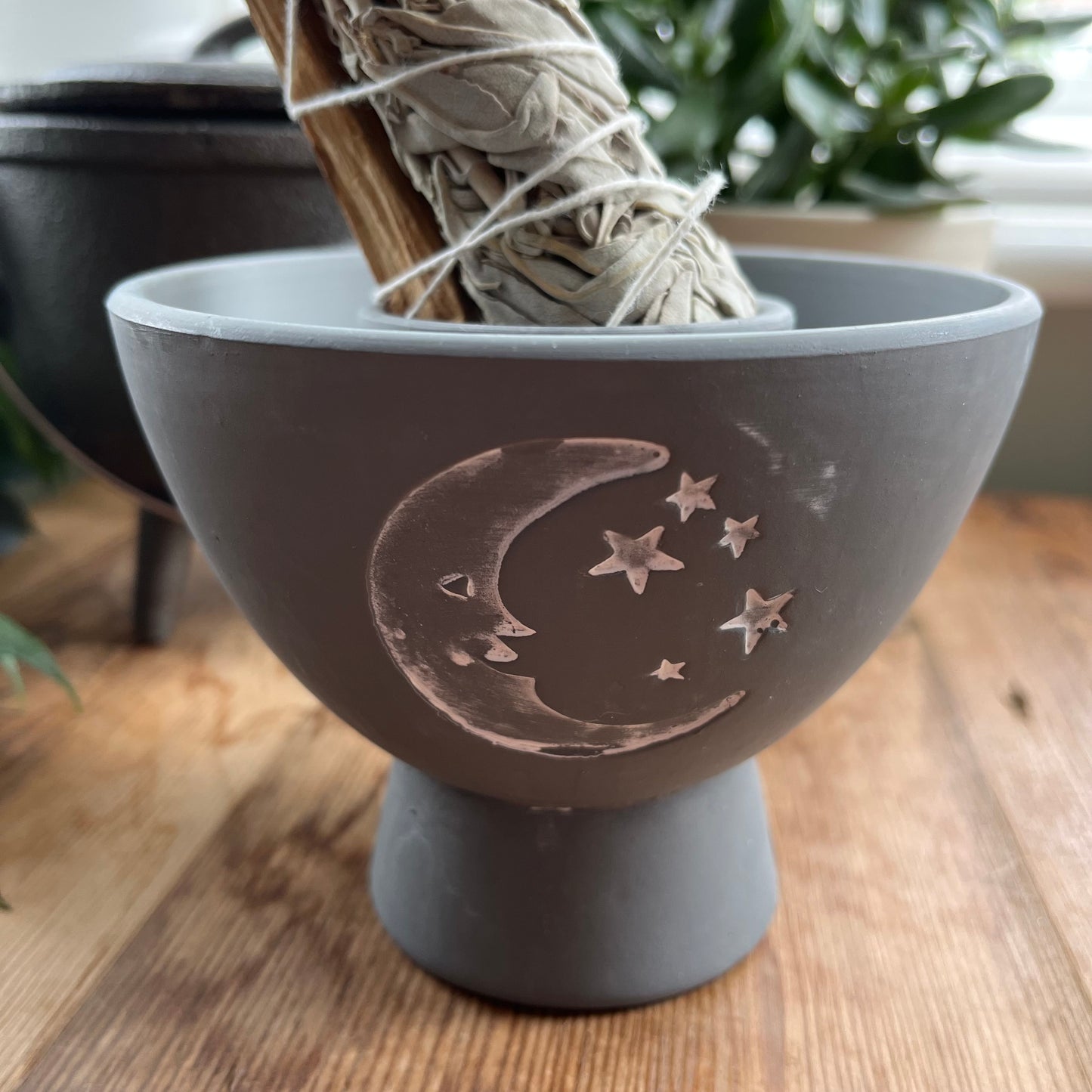 Grey rustic looking pot for holding herbs and smudge sticks while they burn. Has a white painted motif of moon and stars on the side. Photographed with a smudge stick rested in the central purpose made cutout hole designed to hold smudge stick