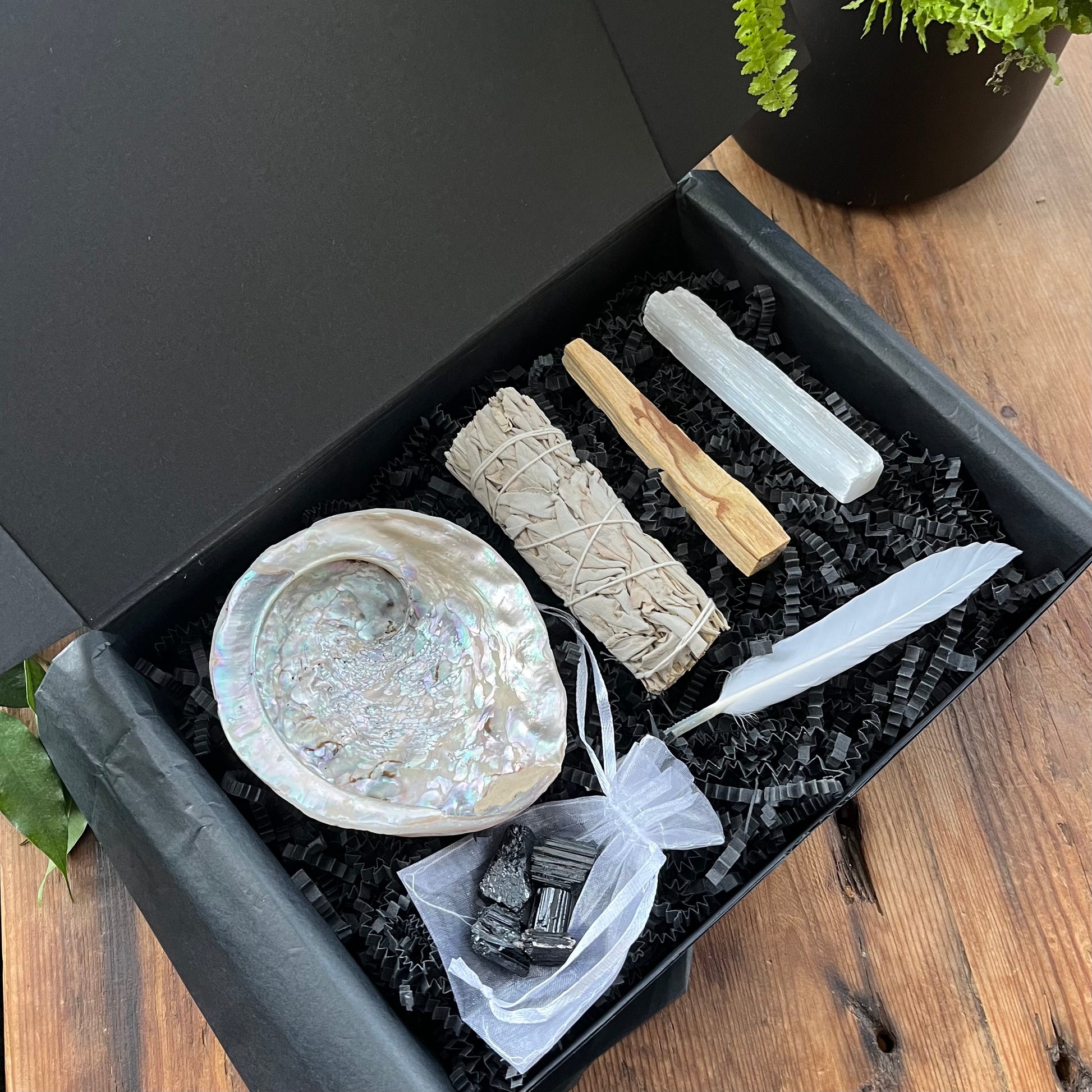 Black gift box exhibiting the contents of our 'sacred cleansing ritual gift box' - abalone shell, sage smoke stick, palo santo stick, rough raw selenite stick, 5 rough black tourmaline gemstones and a white goose feather. phtographed on a wooden table with a plant in the background 