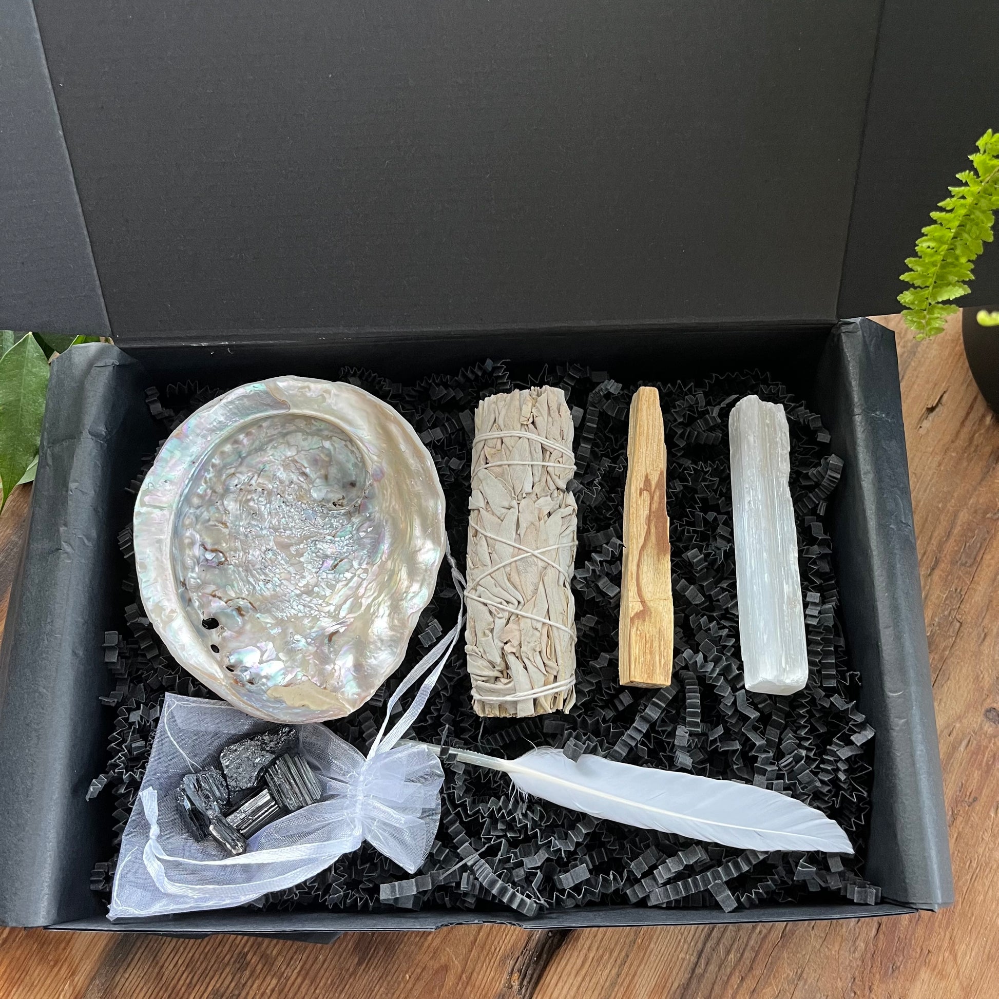 Black gift box exhibiting the contents of our 'sacred cleansing ritual gift box' - abalone shell, sage smoke stick, palo santo stick, rough raw selenite stick, 5 rough black tourmaline gemstones and a white goose feather. phtographed on a wooden table with a plant in the background 