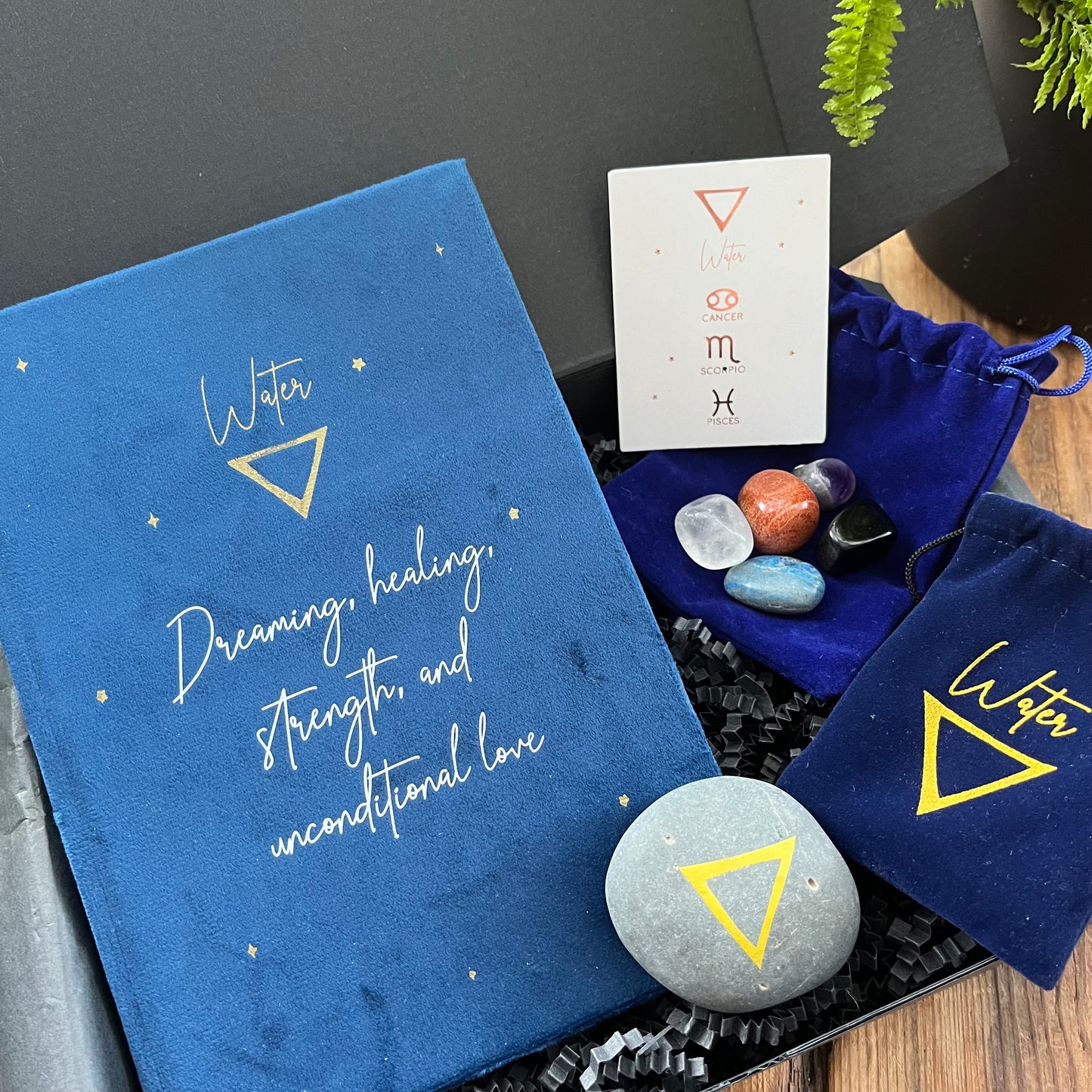 A Black gift box exhibiting the contents of our 'Water signs elemental gift box' - a blue velvet notebook featuring inspiring words and the symbol for water. A grey stone decorated with the elemental symbol for water alongside its matching blue velvet pouch also featuring the same symbol, 5 tumble stones representing water Lapis Lazuli, Black Obsidian, clear Quart, Amethyst and Red Jasper and their pouch and information card. Photographed on a wooden table with a plant in the background.