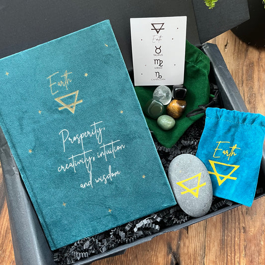 A Black gift box containing our 'earth signs elemental gifts' a Green velvet notebook featuring the elemental symbol for earth and inspiring words, two velvet pouches 1 containing a stone which features the elemental symbol for earth and the other contains tumbled stones associated with the element of earth. Displayed on a wooden table with a plant in the background 