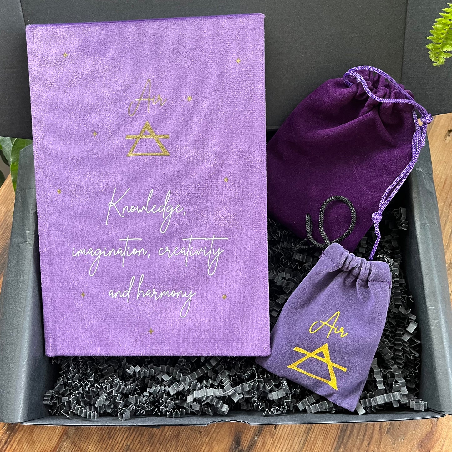 On a wooden table sits the contents of our 'Air Element Gift Box' a purple velvet notebook with inspiring words and the word air and the symbol for air. A grey rune stone decorated with the symbol for air alongside its purple velvet pouch also featuring the symbol for air. 5 tumble stones Lapis Lazuli, Amethyst, tigers eye, fluorite and labradorite, representing the element of air sit alongside their purple velvet pouch. In the back ground is a plant.