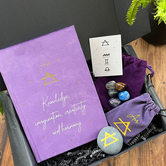 A Black Gift Box containing our ' Air signs elemental gift box' containing a purple velvet notebook with inspiring words and the word air and the symbol for air. A grey rune stone decorated with the symbol for air alongside its purple velvet pouch also featuring the symbol for air. 5 tumble stones Lapis Lazuli, Amethyst, tigers eye, fluorite and labradorite, representing the element of air sit alongside their purple velvet pouch. 