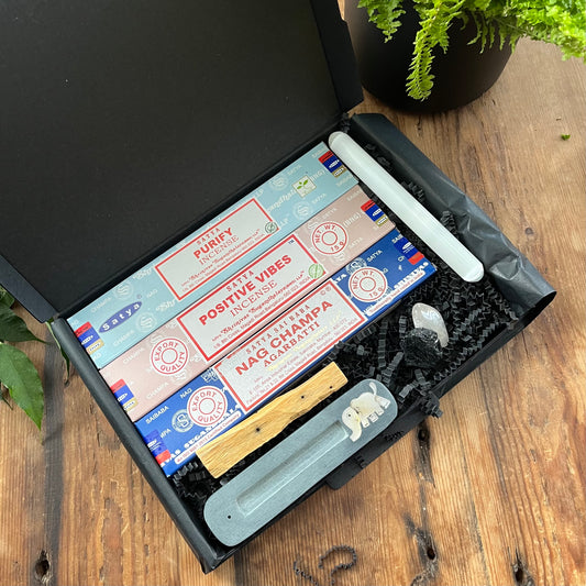 A Black letterbox size gift box containing 3 different boxes of incense Purify,Positive vibes and Nag Champa plus a stone incense holder featuring an elephant inlay, a palo santo wood stick, a smooth selenite wand and 2 crystals 1 unpolished Black Tourmaline and 1 Polished Clear Quartz. Box is pictured on a wooden table with a plant in the background 