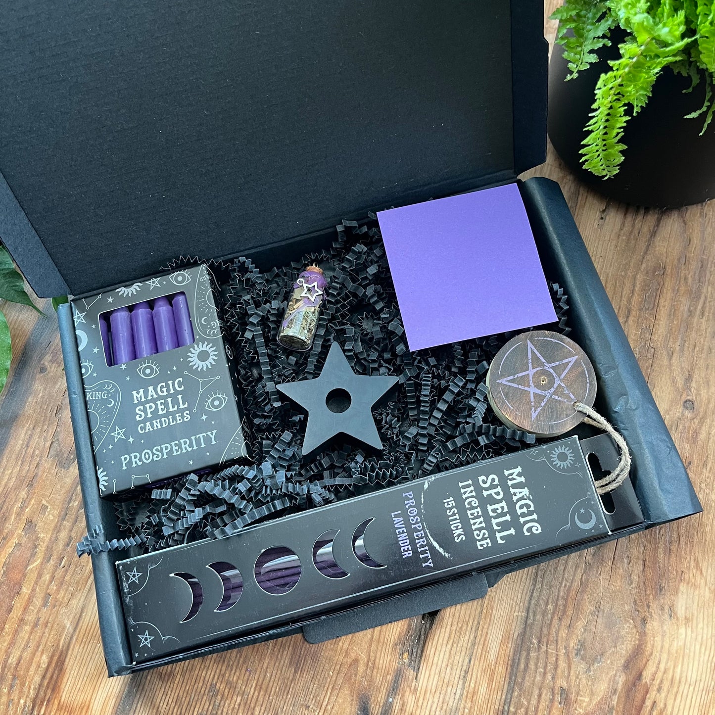 A black letterbox sized gift box containing 'prosperity spell kit''Prosperity Spell Kit' - Pack of lavender scented incense sticks for prosperity, and wooden pentagram incense holder, Pack of purple magic spell candles, wooden black star shaped candle holder, squares of purple paper and a "prosperity' spell bottle containing herbs and sealed with purple wax and a charm. Photographed on a wooden table with a plant in the background 