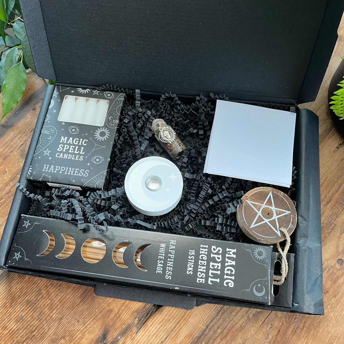 A Black letterbox size box containing the Happiness Spell Kit' Contents - Pack of Happiness white sage Incense sticks, and wooden pentagram incense holder, Pack of white magic spell candles, wooden white spell candle holder with crescent moon and star design, squares of white paper and a happiness spell bottle containing herbs and sealed with white wax and a charm. Photographed on a wooden table with a plant in the background