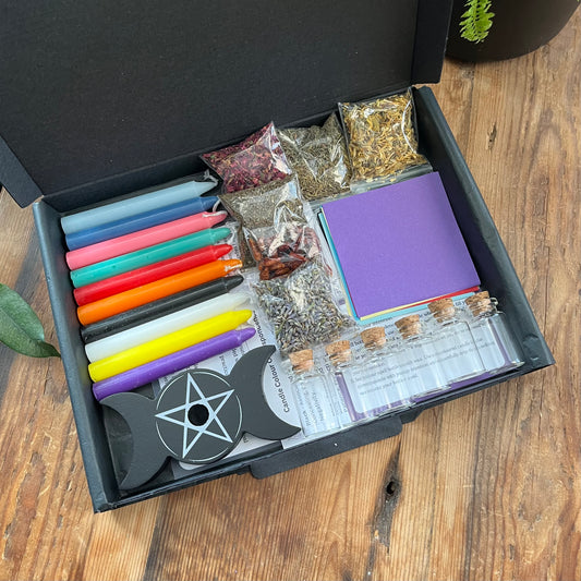 A black letterbox sized gift box cntaining our "candle spell and spell bottle starter kit' - 10 different coloured candles, a spell candle holder, coloured paper squares to match the candles, 6 small spell bottles, 6 clear plastic packets filled with a variety of herbs, spies and flowers and instruction sheets. Photographed on a. wooden table with a plant in the background. 