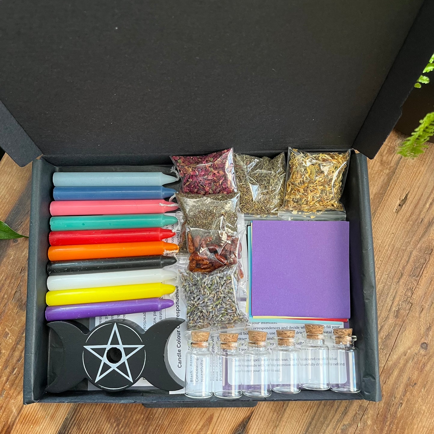 A black letterbox sized gift box cntaining our "candle spell and spell bottle starter kit' - 10 different coloured candles, a spell candle holder, coloured paper squares to match the candles, 6 small spell bottles, 6 clear plastic packets filled with a variety of herbs, spies and flowers and instruction sheets. Photographed on a. wooden table with a plant in the background. 