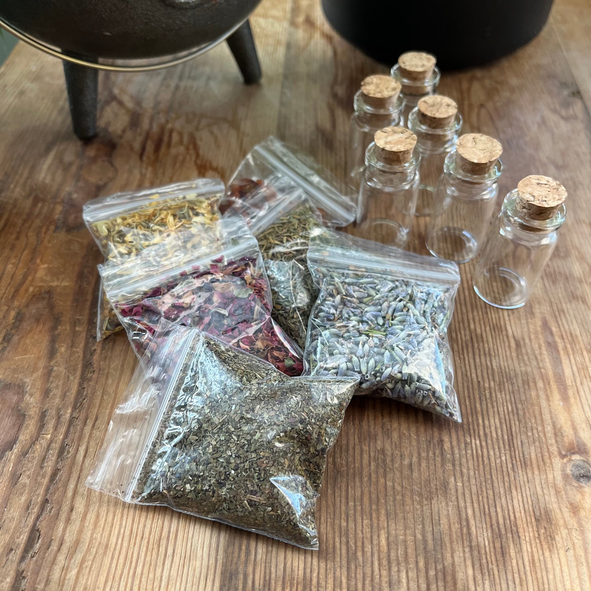 6 empty spell bottles sit on a wooden table next to packets of herbs, spices and dried flowers. 