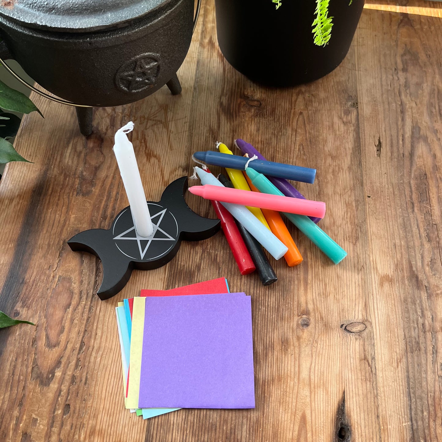 10 coloured spell candles, a spell candle holder in the shape of a triple moon with a pentagram design and a pile of coloured paper squares. sit on a wooden table next to a cauldron ready for spell work. 