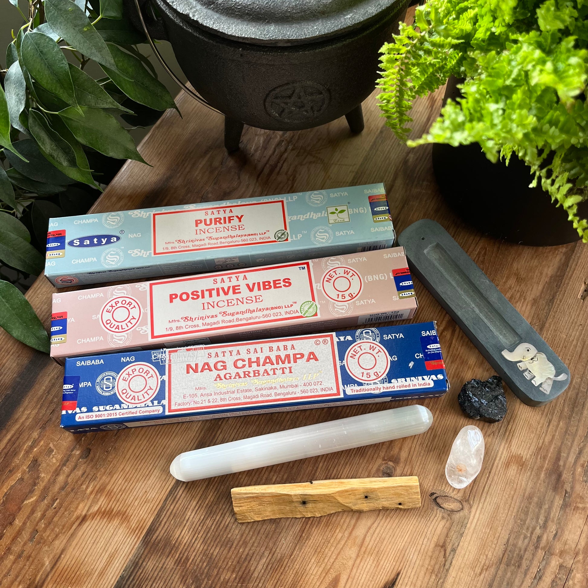 The contents of the Sacred serenity  gift box - 3 different boxes of incense Purify,Positive vibes and Nag Champa plus a stone incense holder featuring an elephant inlay, a palo santo wood stick, a smooth selenite wand and 2 crystals 1 unpolished Black Tourmaline and 1 Polished Clear Quartz. Contents are pictured on a wooden table with a plant in the background 